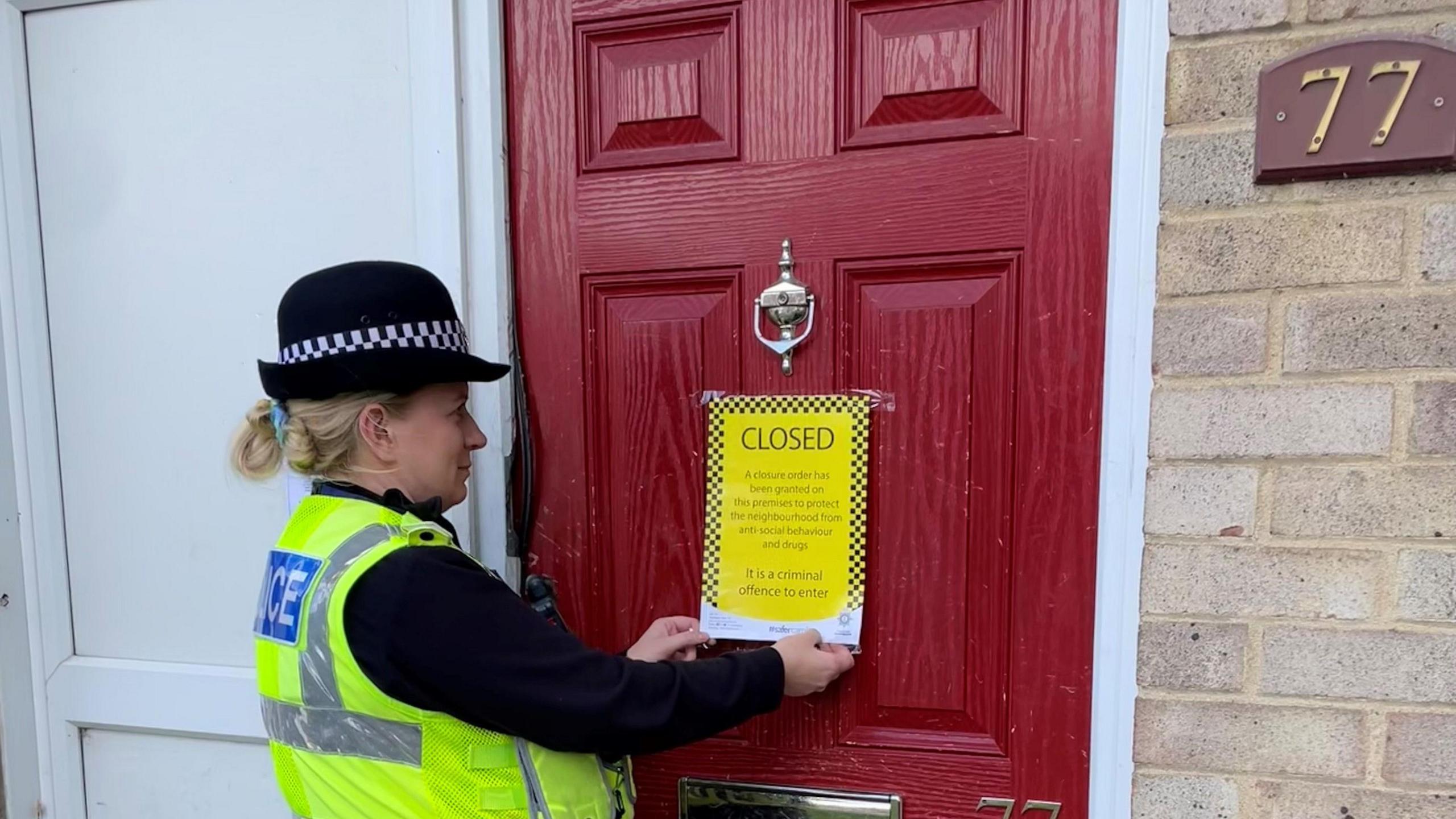 A police officer smiles as she hangs a yellow closure order on a red front door of a property. To the right of the door is the number 77.