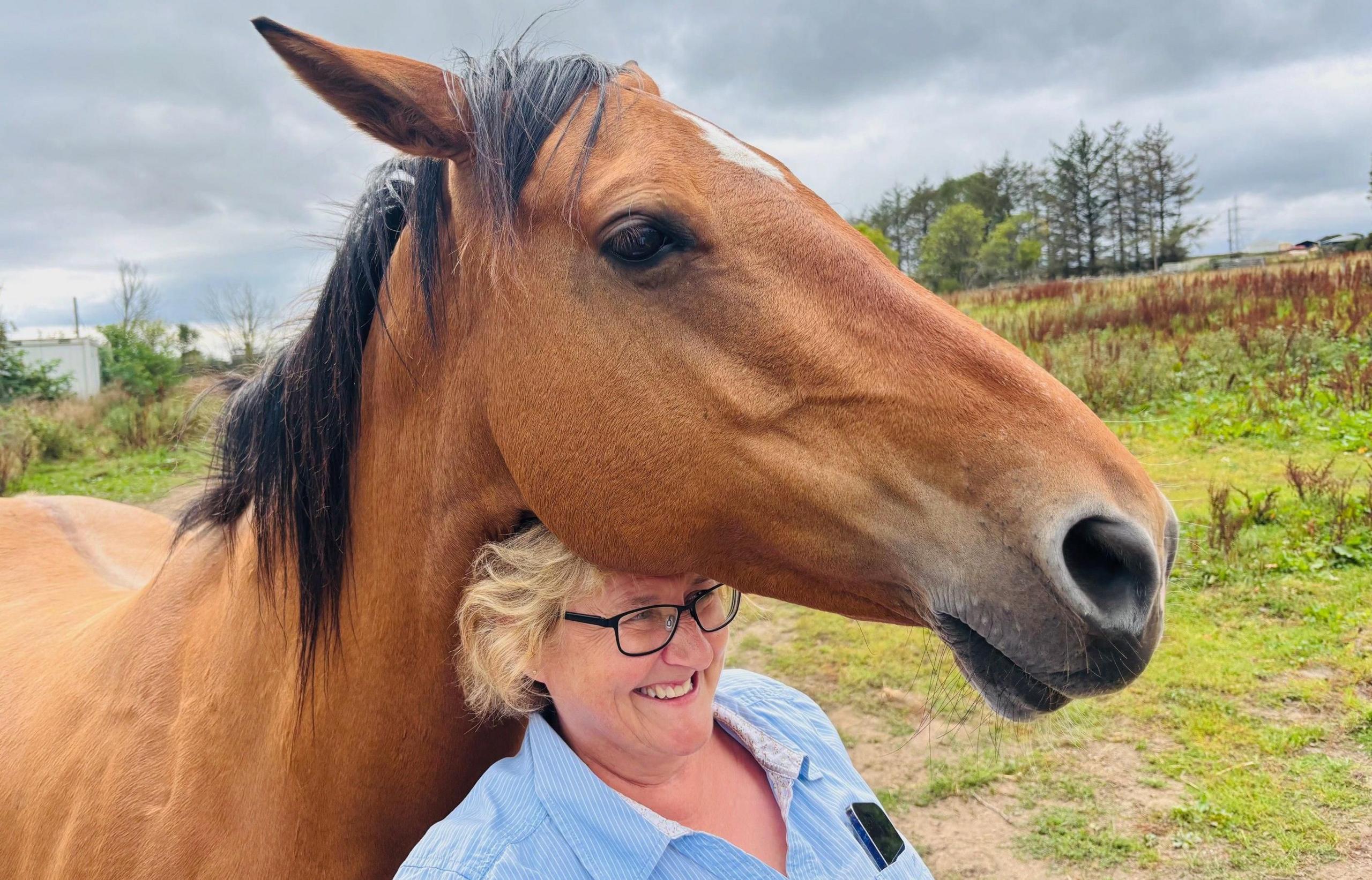 Louise Yule, smiling, with her head under a brown horse's face, in a field.