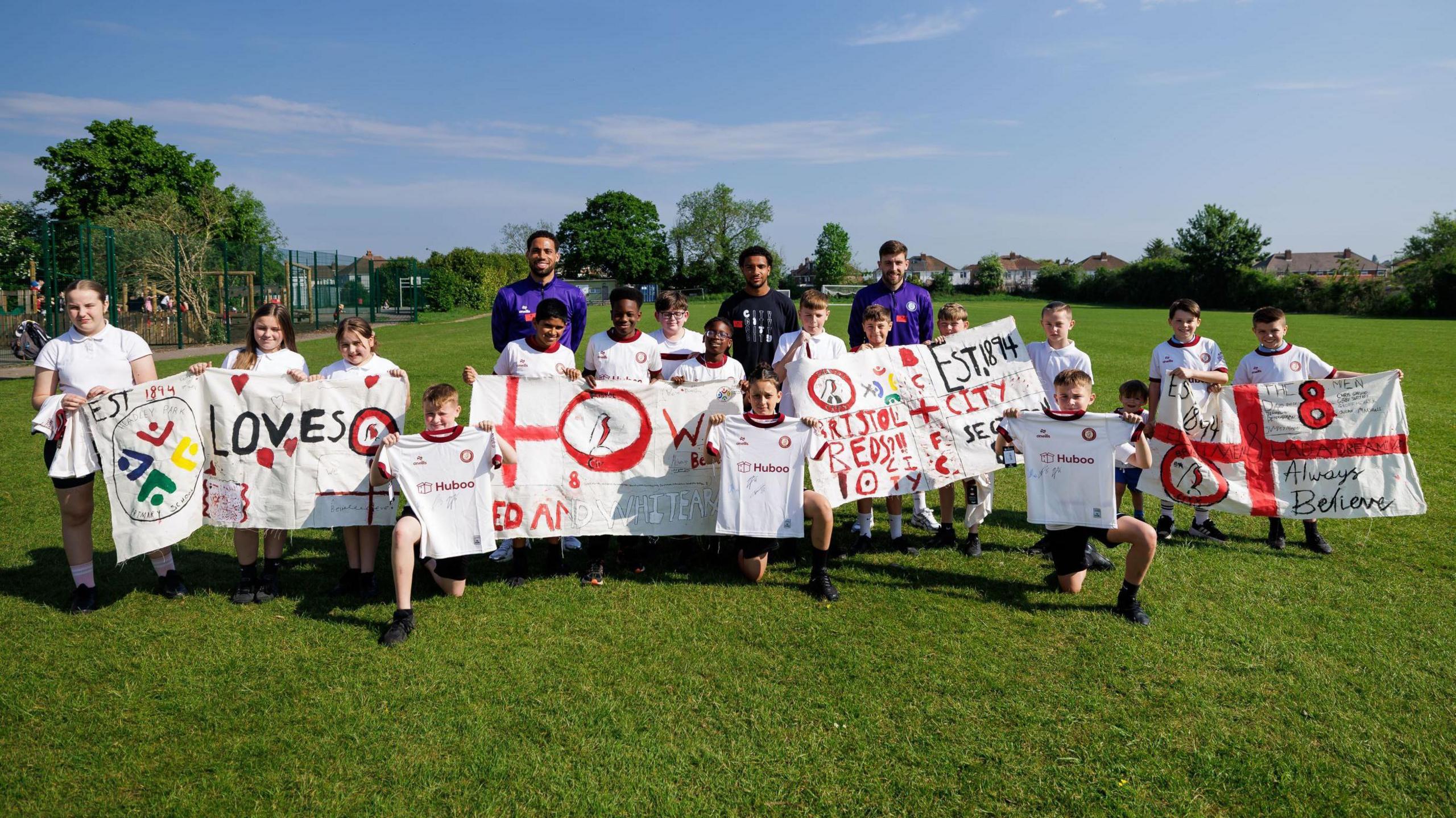 A group of school pupils at Headley Park Primary School hold up several banners showing their support for Bristol City ahead of their game against Preston. Standing behind the children, who are on the school's playing field, are City players Zak Vyner, Joe Williams and Elijah Morrison.
