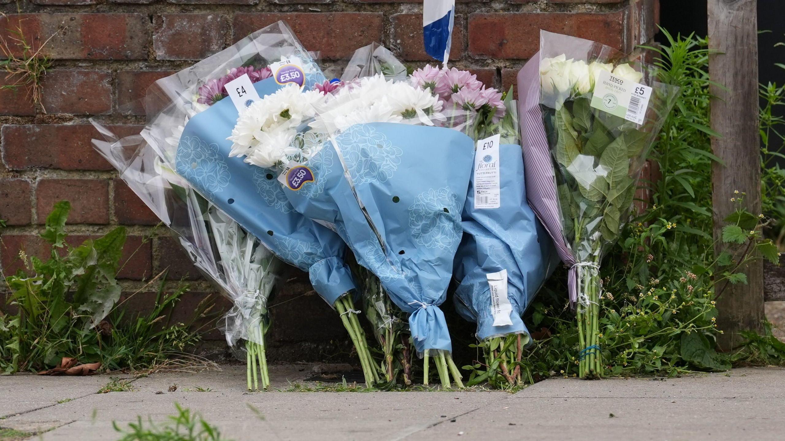 Six bunches of flowers in plastic packaging leant against a brick wall on a pavement.