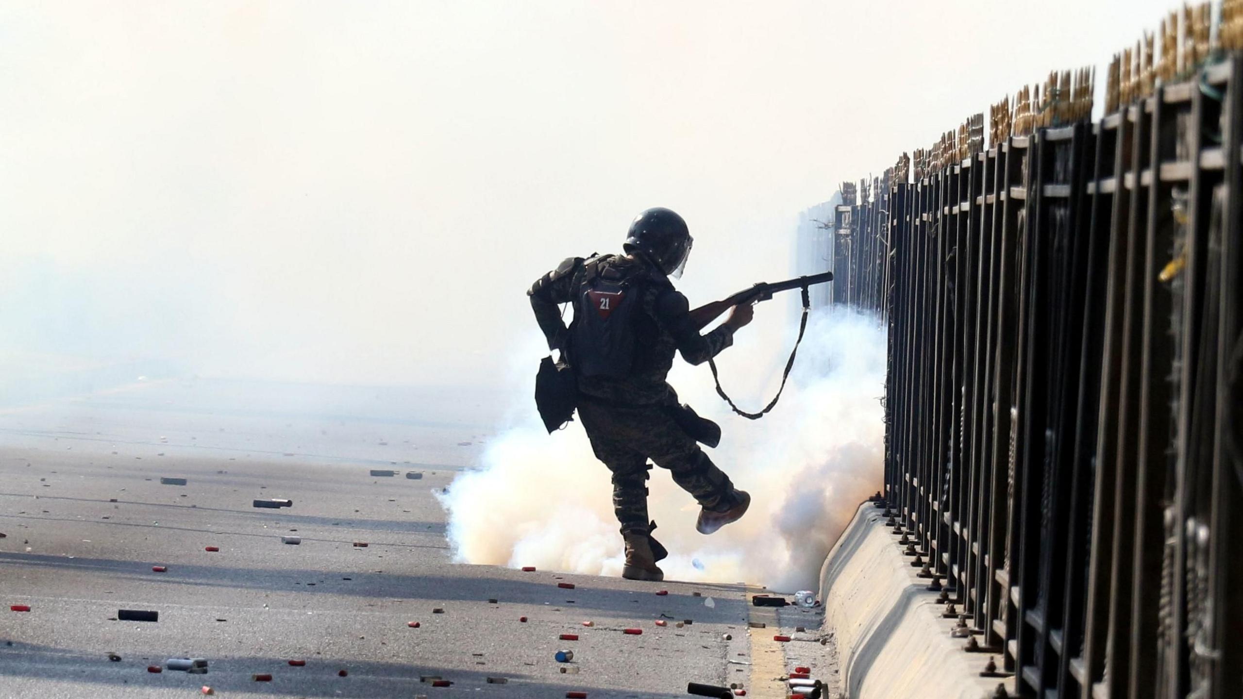 A soldier appears to fire tear gas on a street littered with cartridges. He is silhouetted against the smoke