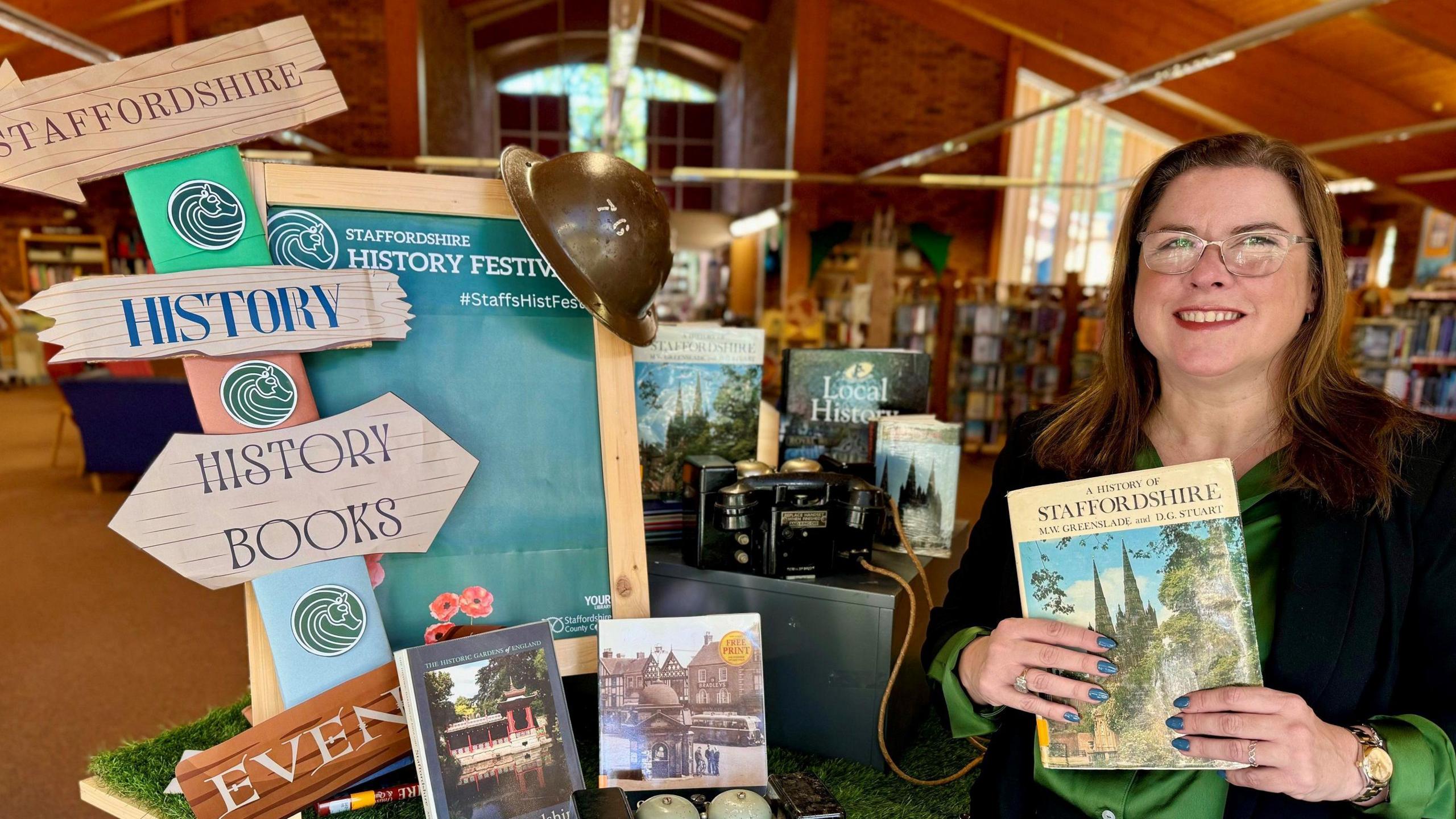 A woman holds up a history book about Staffordshire whilst standing next to a display to promote the forthcoming history open day.