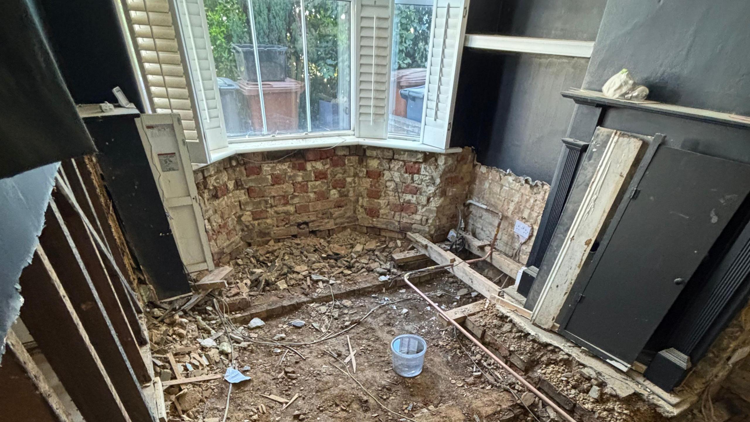 One of the rooms in Jay's house with a bay window, with plantation style shutters open. The floor has been stripped back to soil and joist level. Exposed pipes can be seen alongside debris. There is a black radiator on the right wall.