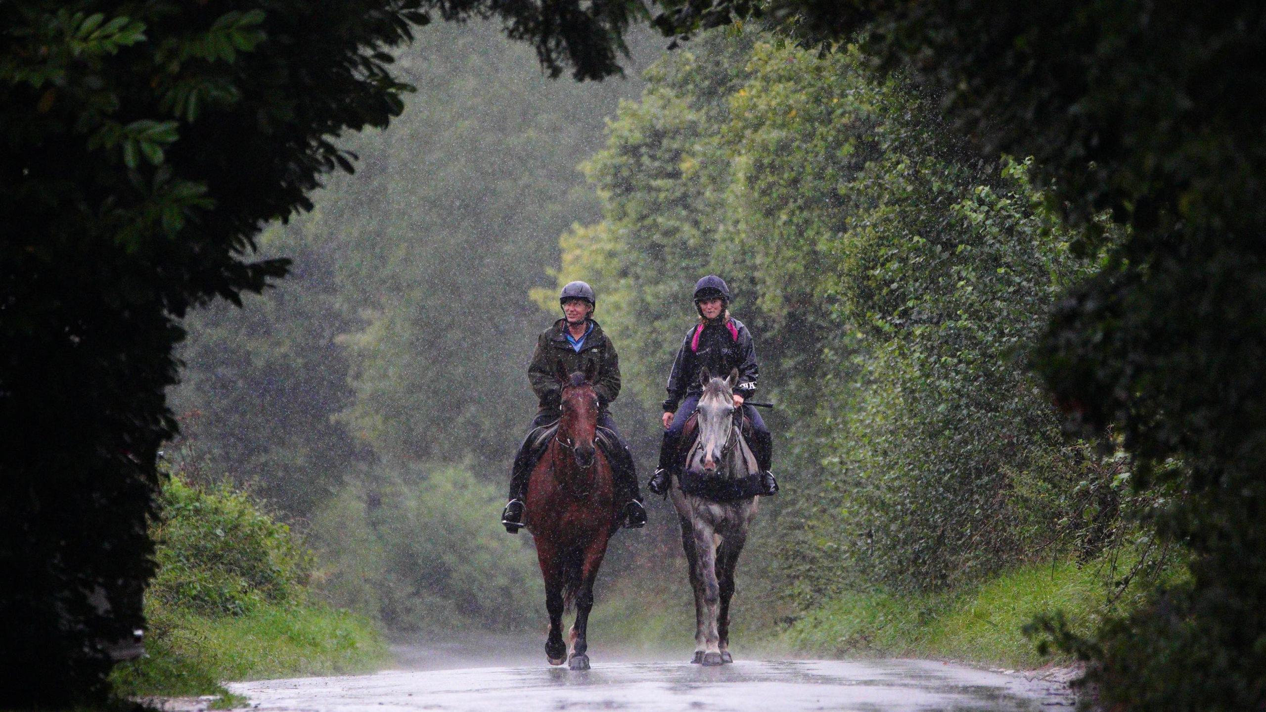 Two female horse riders are seen walking their horses towards the camera. It is raining heavily and the riders are framed by dark foilage