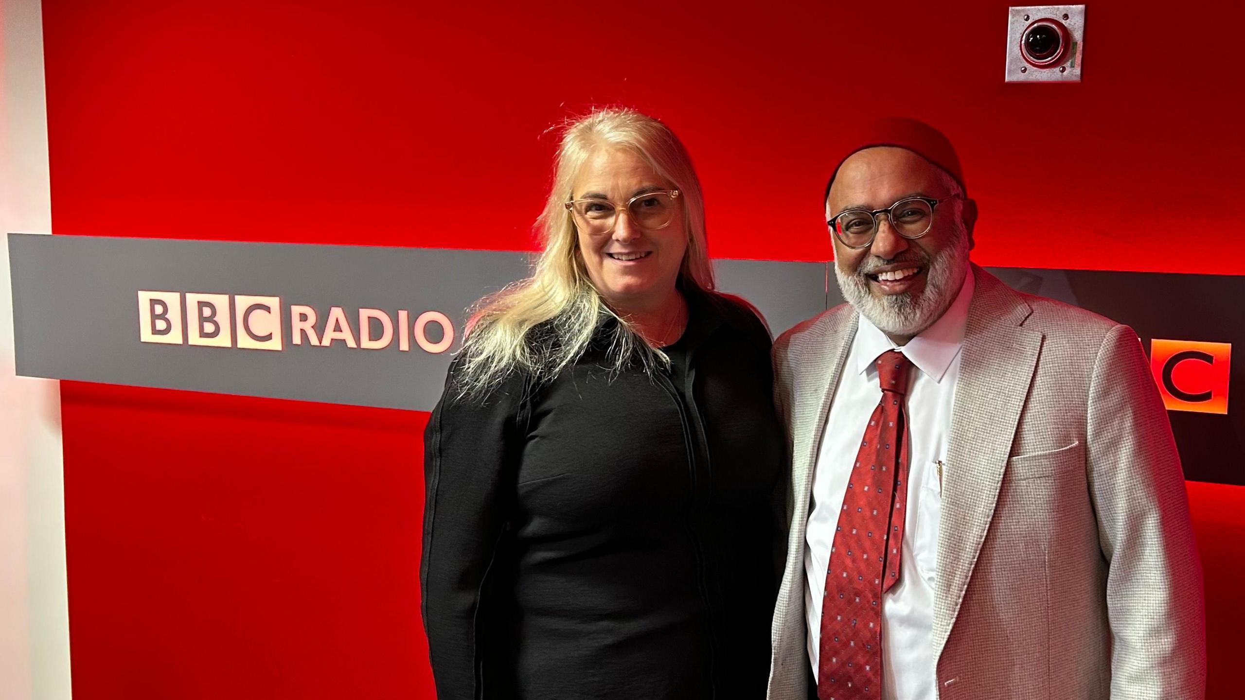 Selina Sasse and Imam Monawar Hussain smiling for the camera at BBC Oxford's radio studio. The logo is on the wall behind them.