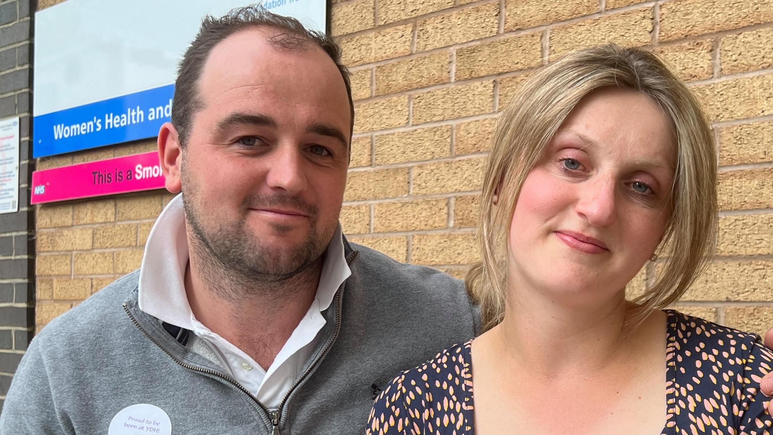 A man wearing a grey half zip jumper looking at the camera with a woman beside him