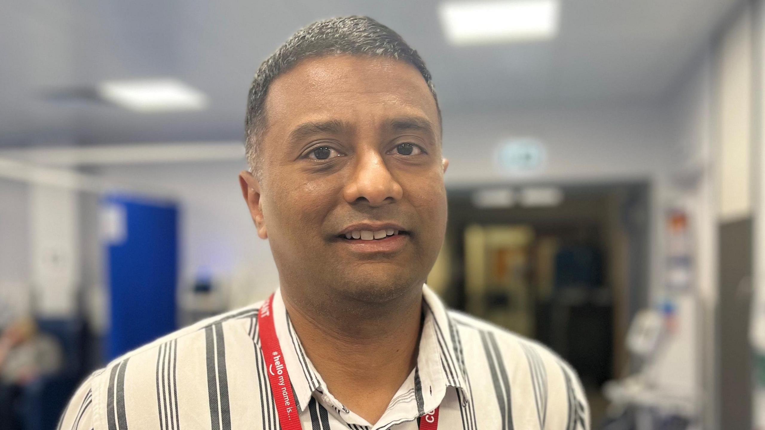 Dr Darshan Kumar is looking directly at the camera. He is wearing a striped white and grey shirt. The background is blurred but appears to be a clinical setting. He is wearing a red lanyard around his neck.