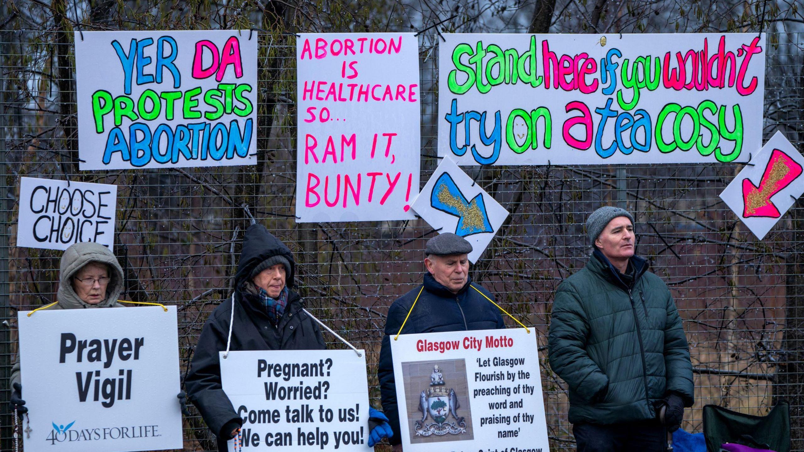 Four protestors standing by a chain link fence, holding signs saying things like 'pregnant? worried? Come talk to us!" and £prayer vigil". Pinned to the fence behind them are signs saying things like "yer da protests abortion" and "abortion is healthcare so ram it bunty" 