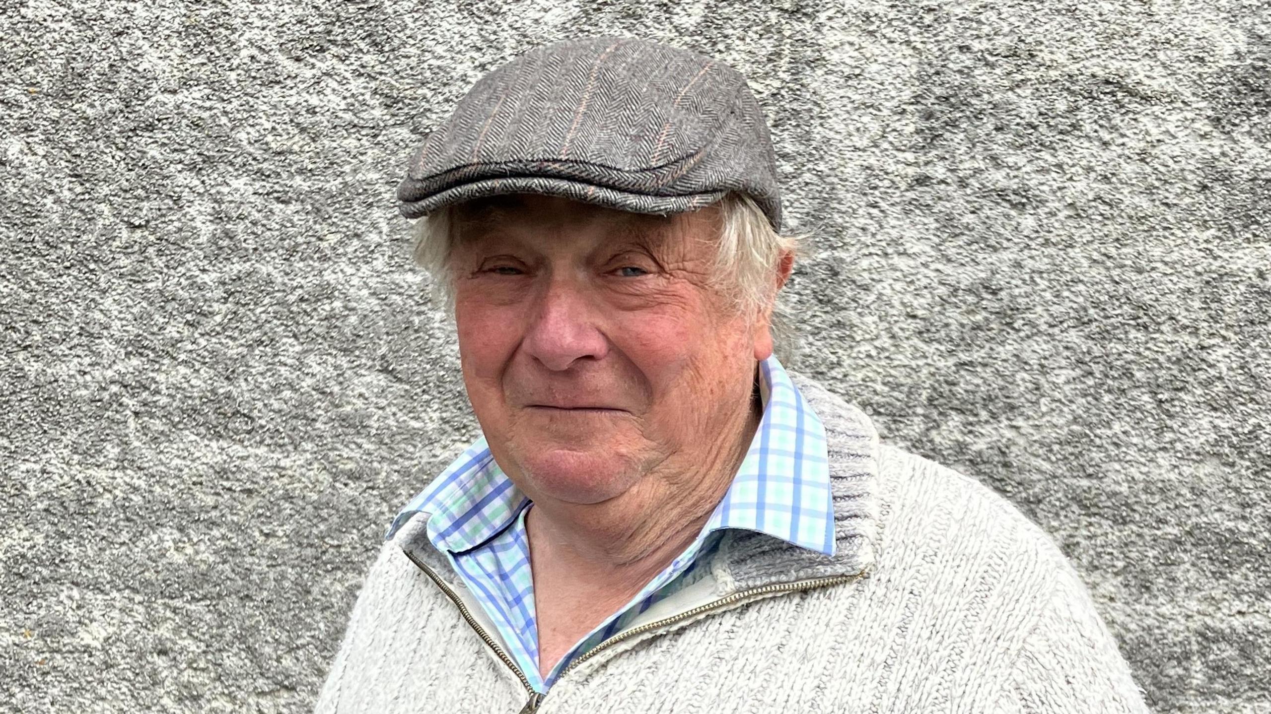 A man with grey hair wearing a grey flatcap, white blue chequed shirt and cream woolen quarter-zip jumper standing in front of a grey stone wall. 