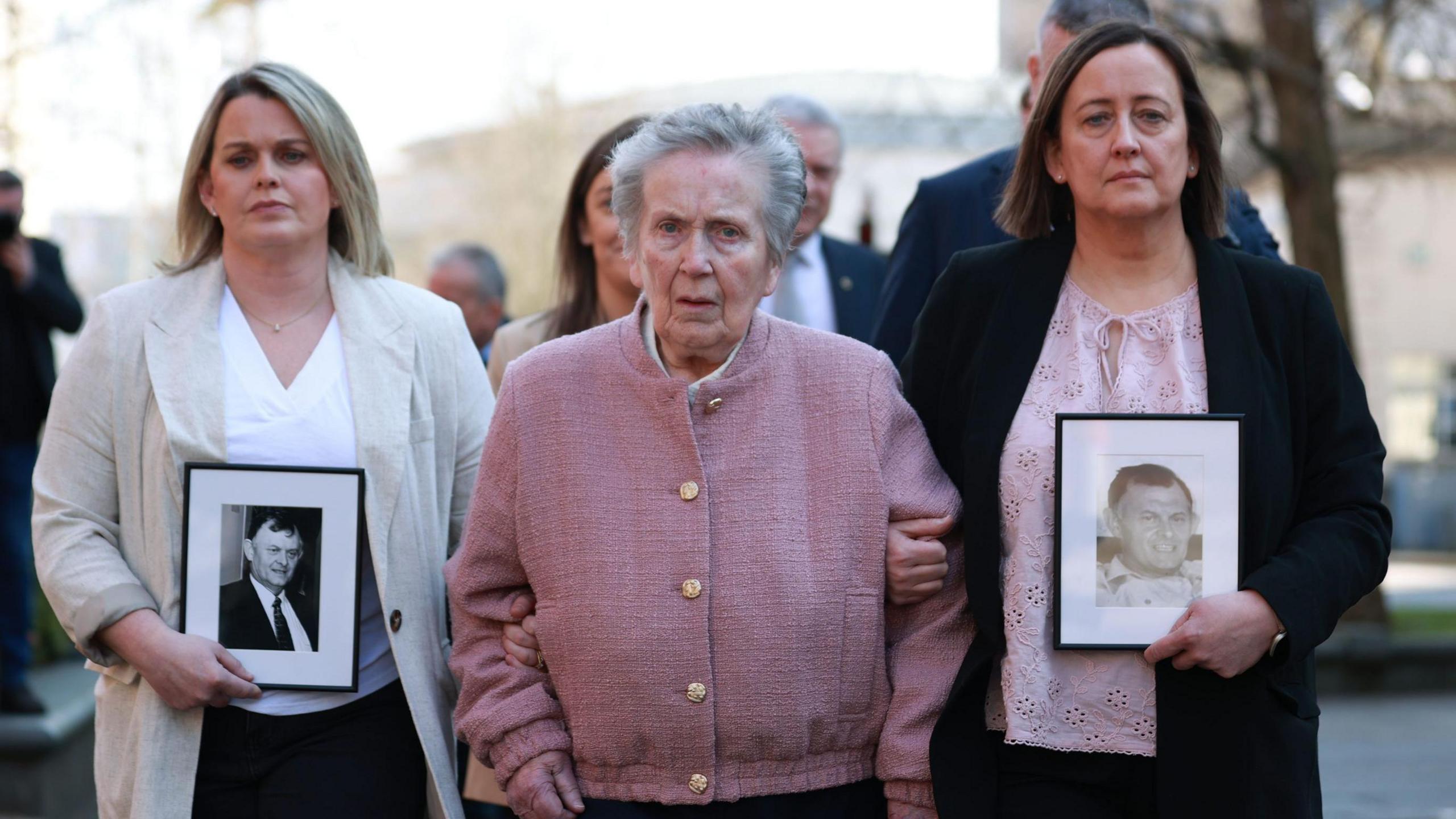 Sean Brown's widow Bridie wearing a pink cardigan with gold buttons walks along flanked by her daughters Siobhan and Claire, who are both holding black and white photos of Sean Brown. Siobhan is wearing a linen jacket over a white top and black trousers, Claire is wearing a dark jacket over a pink top and black trousers.