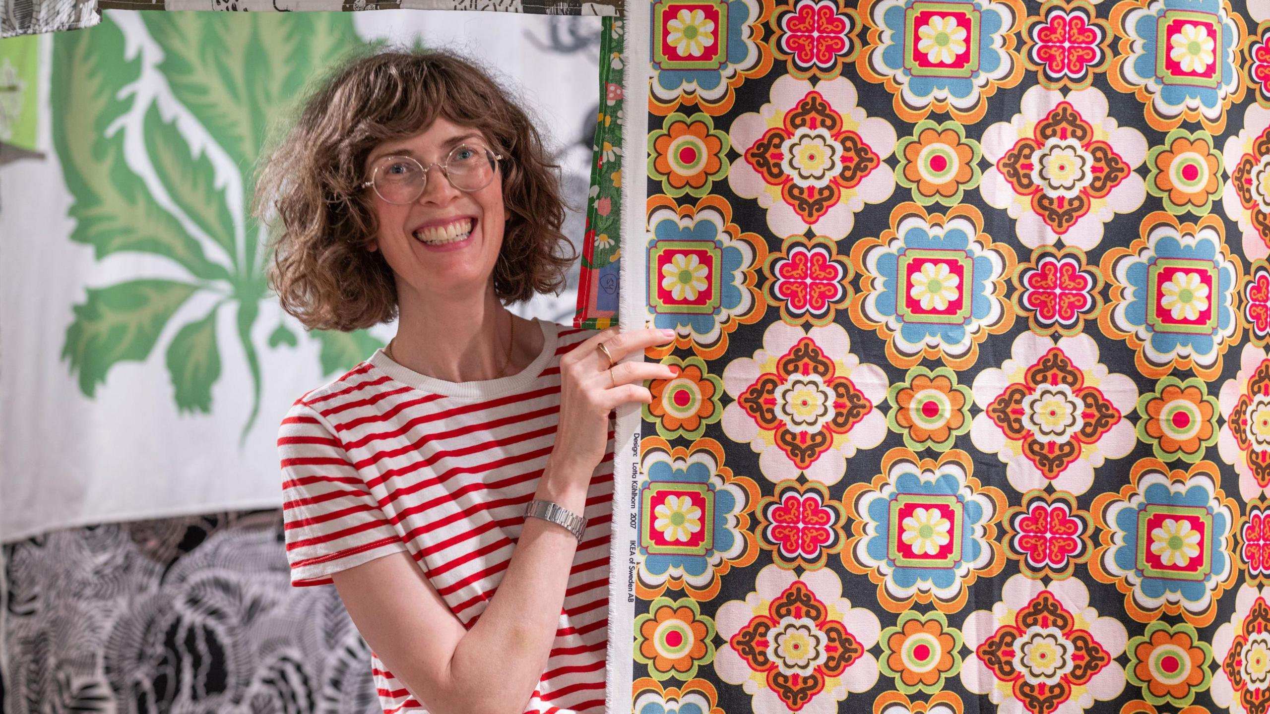 Anna Sandberg Falk, a smiley woman with brown curly hair, peeks out from behind a colourful tile-patterned panel of fabric - several different bold designs hang behind her.