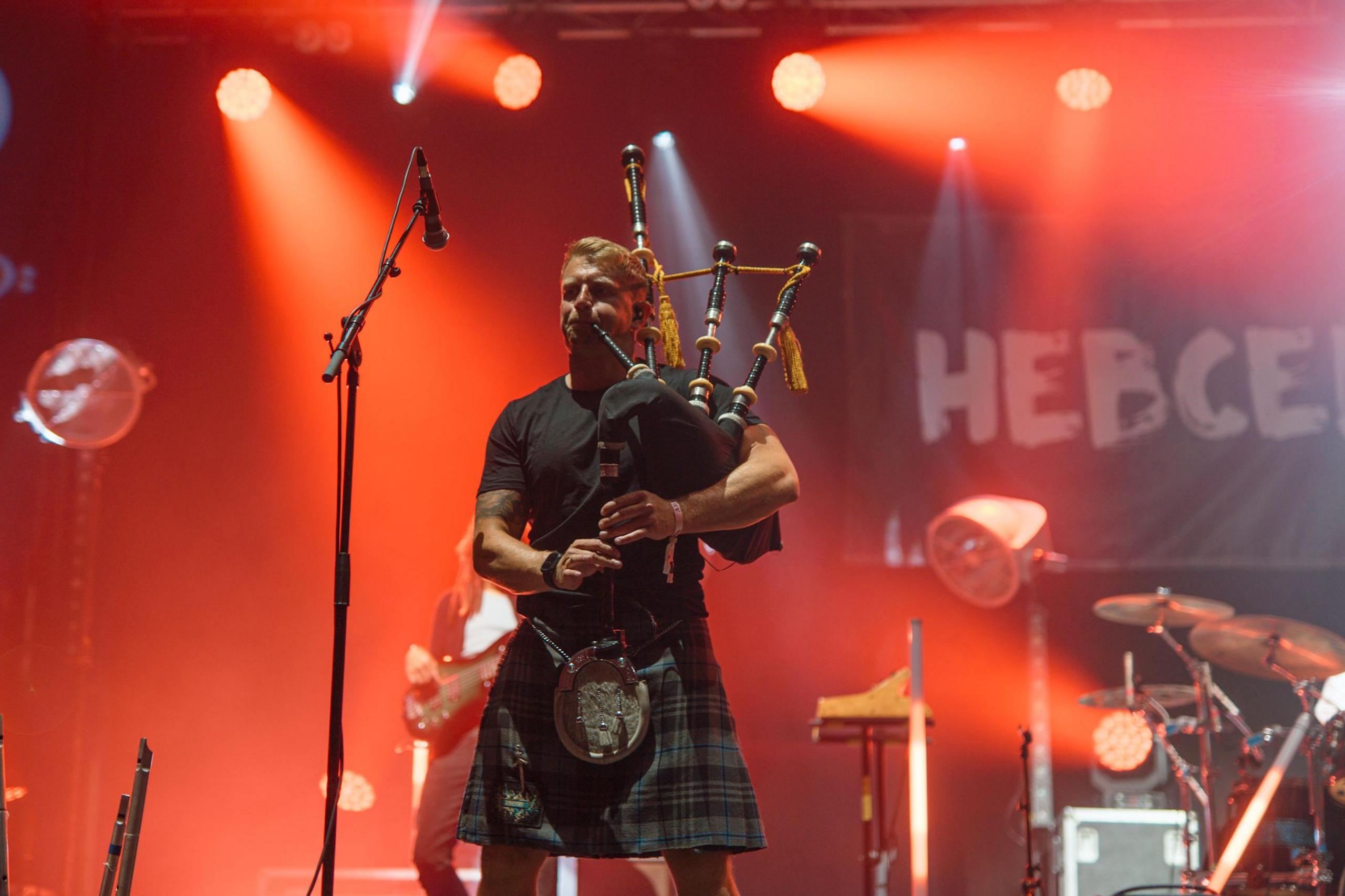 Skerryvore piper play in a kilt. The stage is lit up in red and there are microphones and drums on it