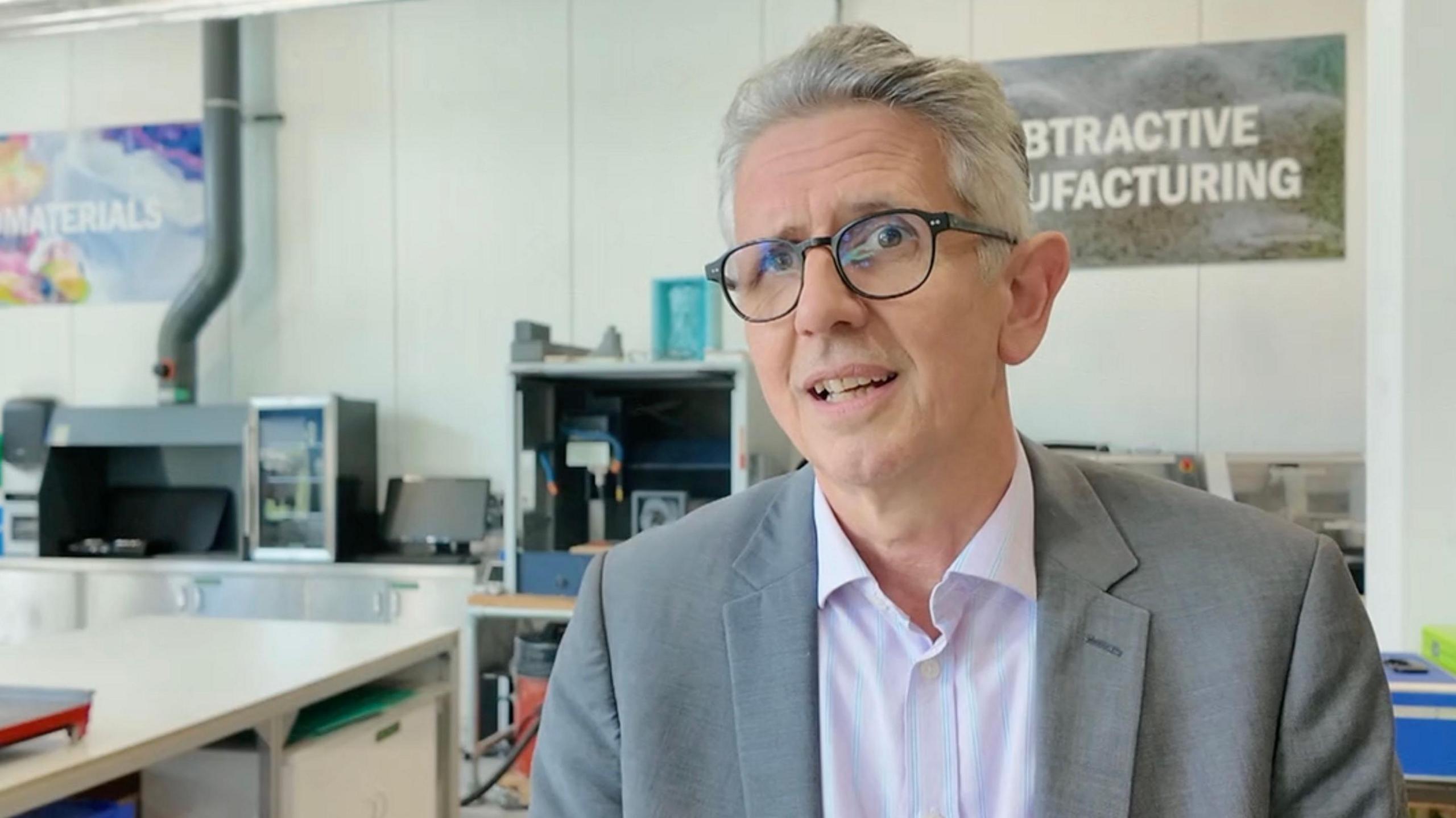 A picture of Stephen. He has grey hair, glasses, is wearing a pink shirt and has a grey blazer. He is pictured in front of an office with machinery and signs on show.