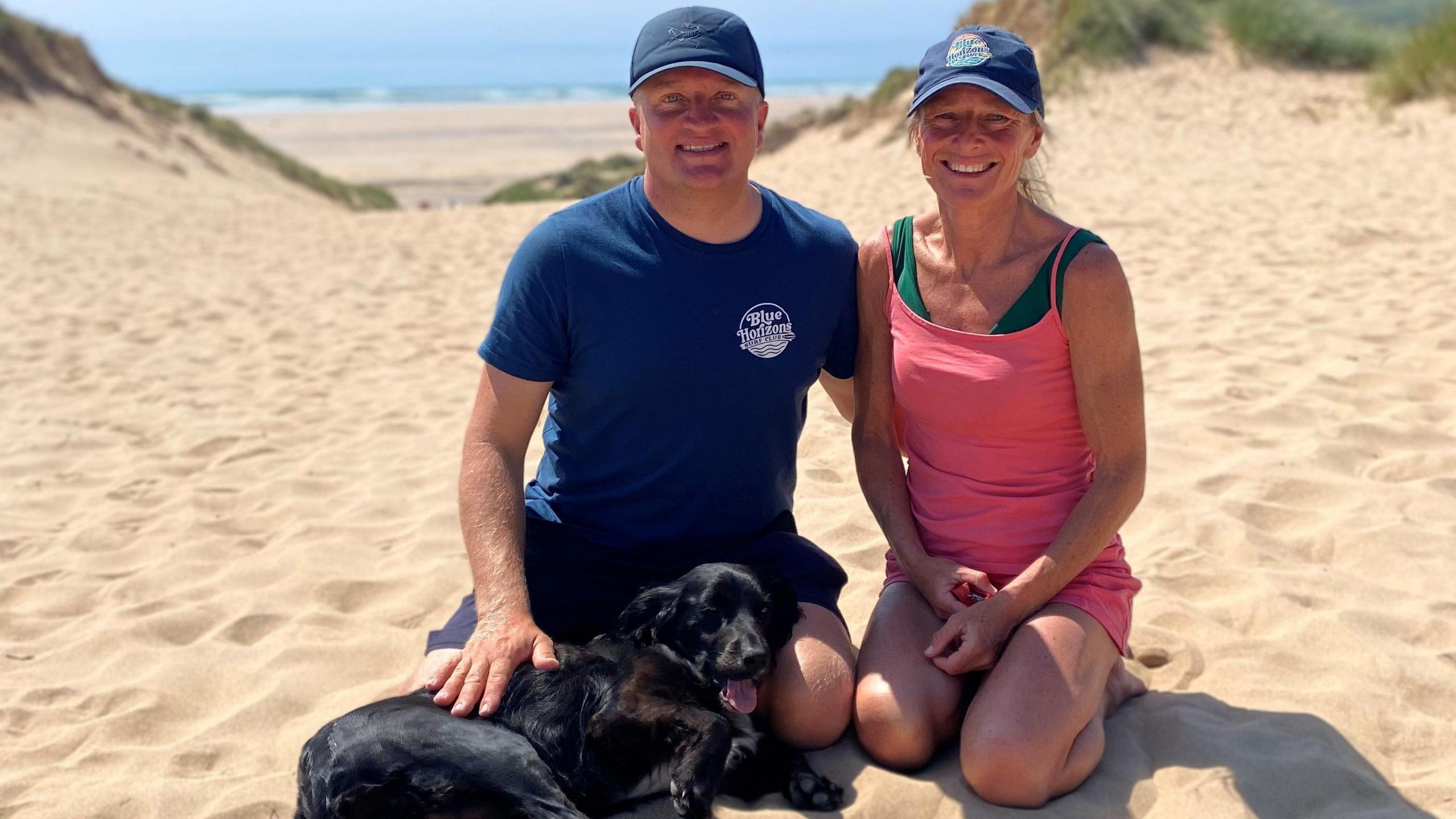 Ariel, the formerly six-legged dog, lying on a beach alongside her owners who are kneeling