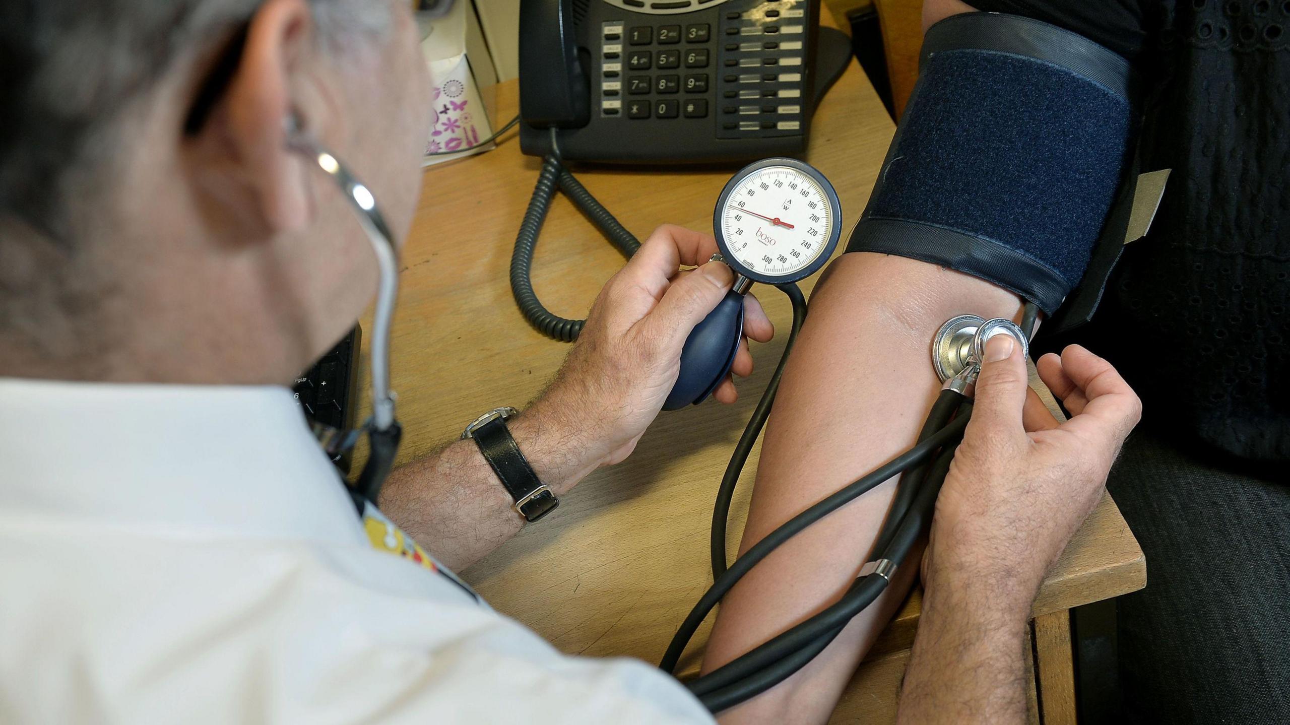 A doctor checking a patient's blood pressure with their arm leaning on a desk with a telephone on it.