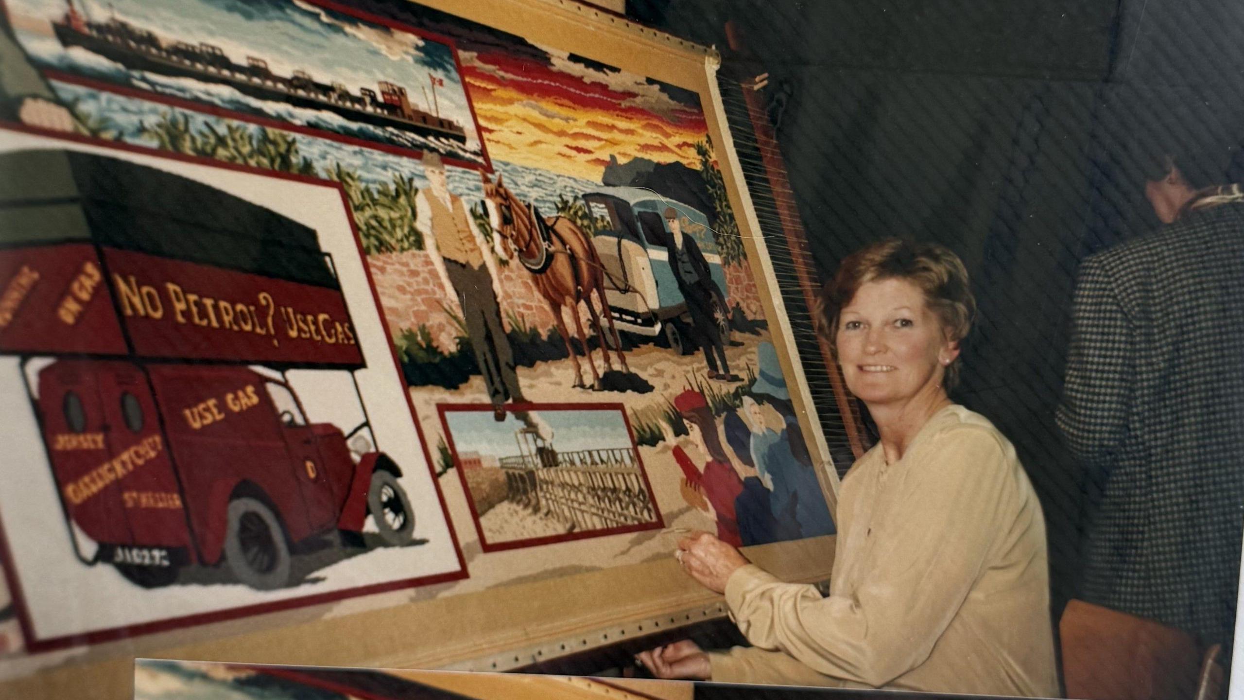 The picture shows a large, colourful textile artwork mounted on a wooden frame. The artwork appears to be a detailed tapestry panel depicting scenes from Jersey’s history, including a red vintage delivery van with the words “No Petrol? Use Gas” and “Jersey Gas” clearly visible, a horse pulling a cart in a rural setting, a background featuring fields, fences, and a vivid sunset sky with shades of orange, yellow, and purple.