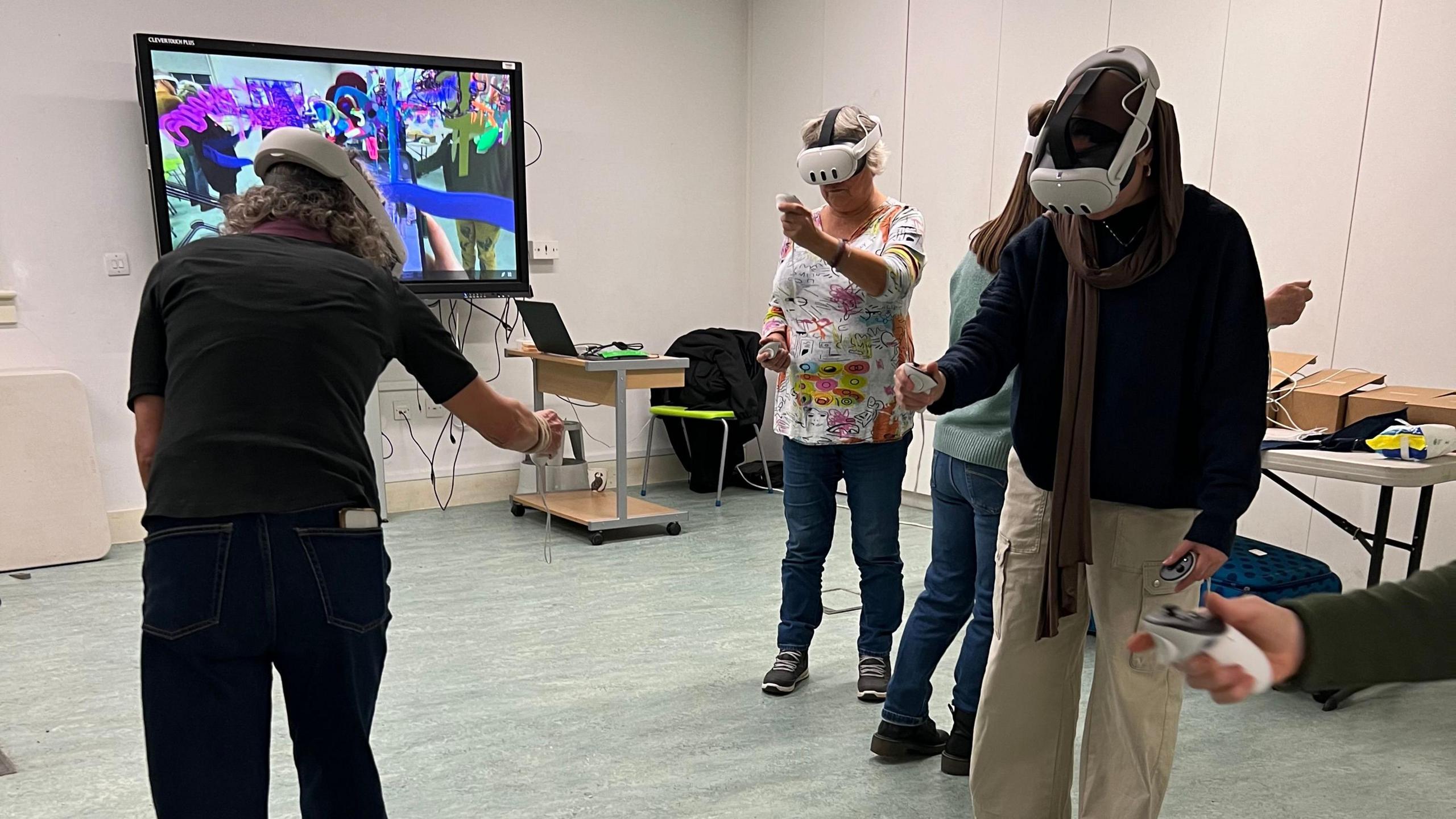 A group of people creating artwork using virtual technology, they are wearing headsets and have a virtual paint brush. They are in a room with a monitor, a laptop, desk, tabled and  chairs. 