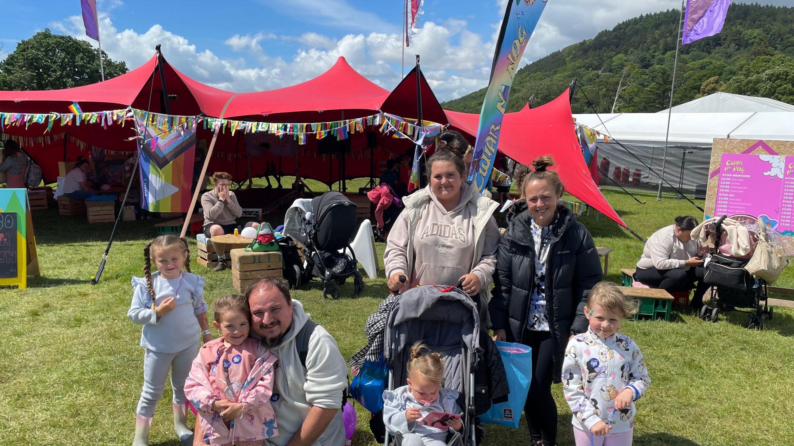 Two women and a man stand with their four children. One sits in a pram, that a woman in a white hoody and coat is holding, and the rest stand and smile at the camera. They stand in front of a red tent. People can be seen sat on the grass behind them and a row of colourful bunting in the background.