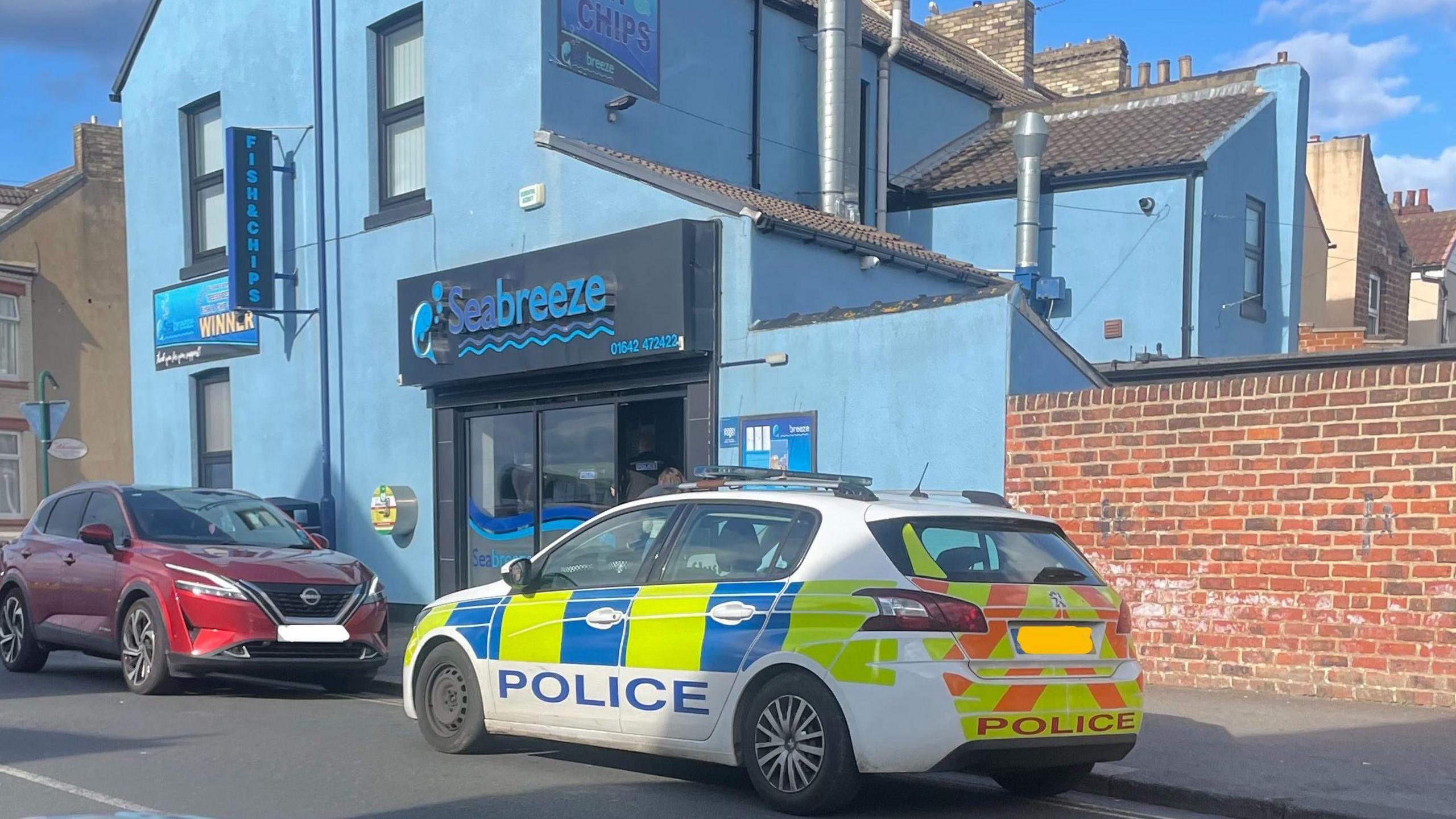 A police car parked outside a blue building with a fish and chip shop called Seabreeze in the centre. The police car is parked on double yellow lines. 