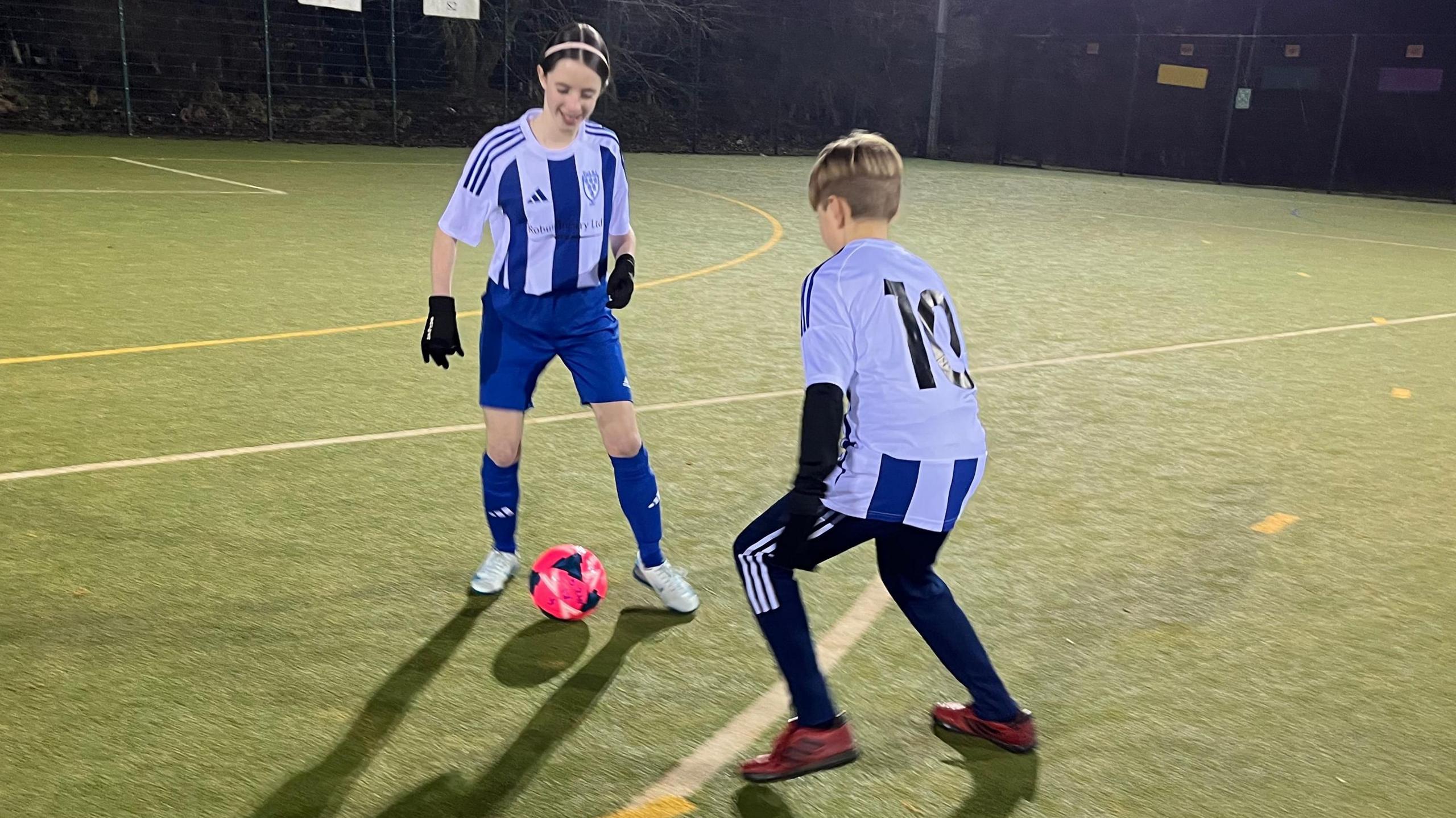 Two girls wearing blue and white football kits are playing football with a pink ball on a floodlit pitch.
The girl nearest the camera has short hair, the other girl has her hair tied back. 