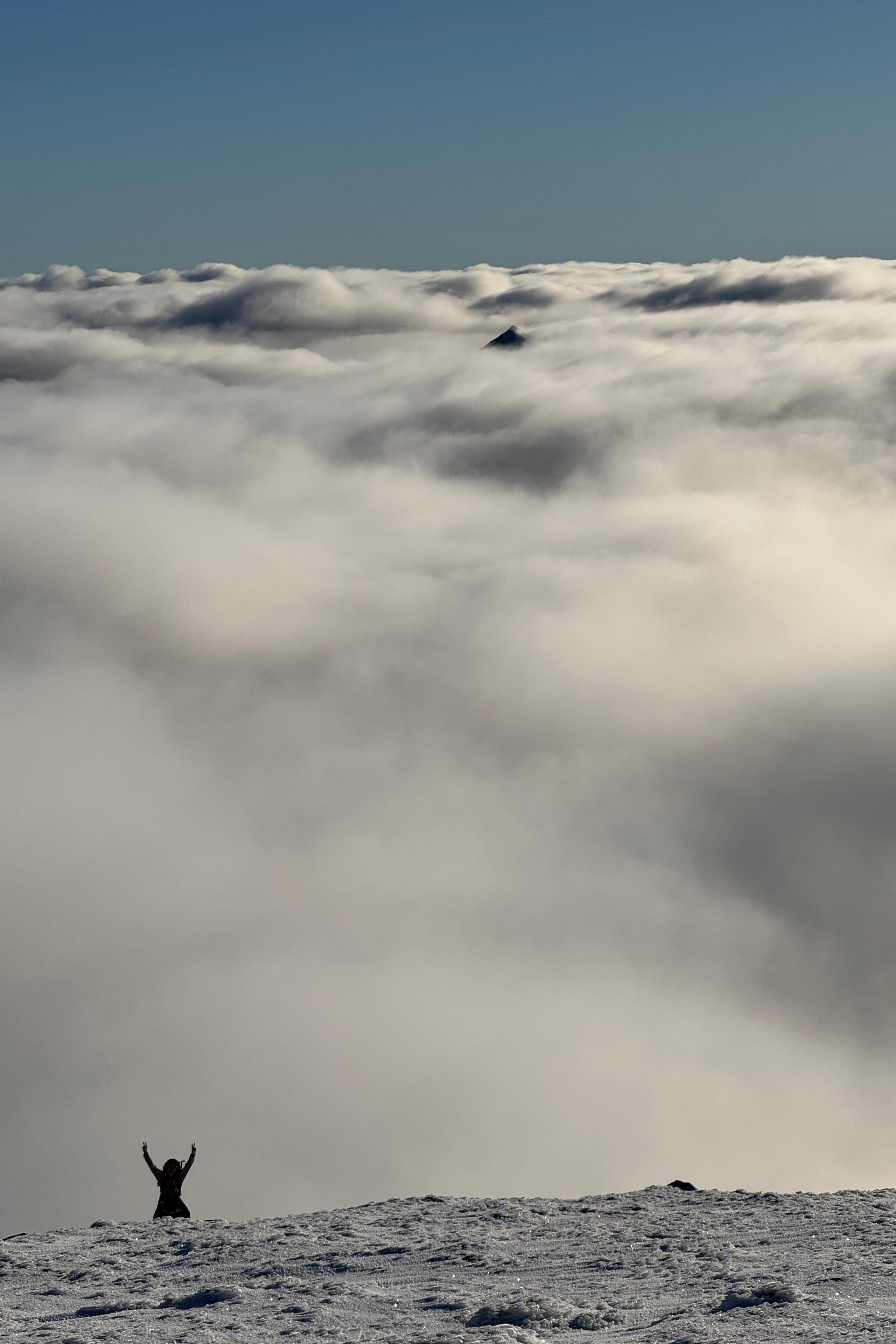 The silhouette of a person, appears small, in front of the clouds and standing on snow