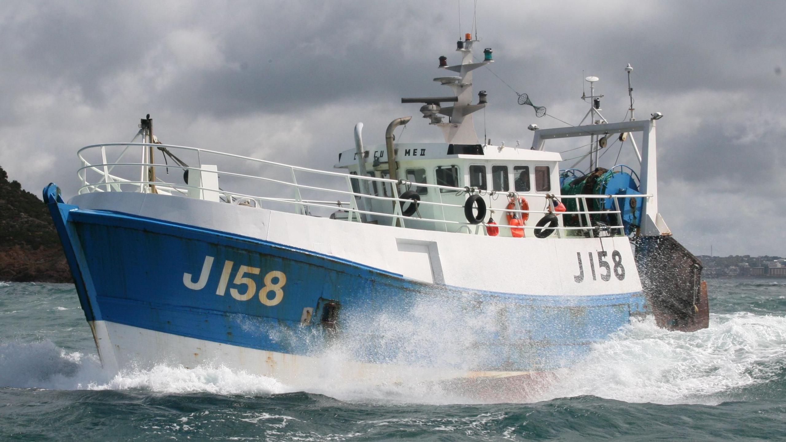 A white and blue fishing boat with J158 on the left side, in the ocean, next to another vessel out at sea with grey skies and land seen in the background.
