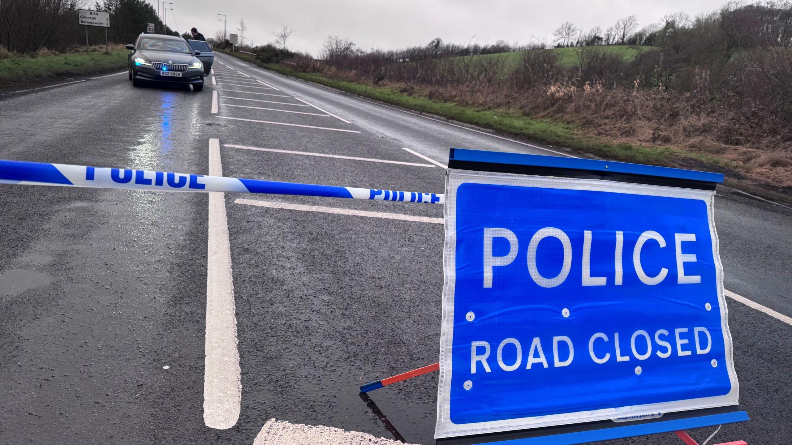 In the middle of the road a line of blue police tape runs across and behind a blue police road sign that reads 'Police Road Closed.' Beyond the tape is a black unmarked Skoda police car.