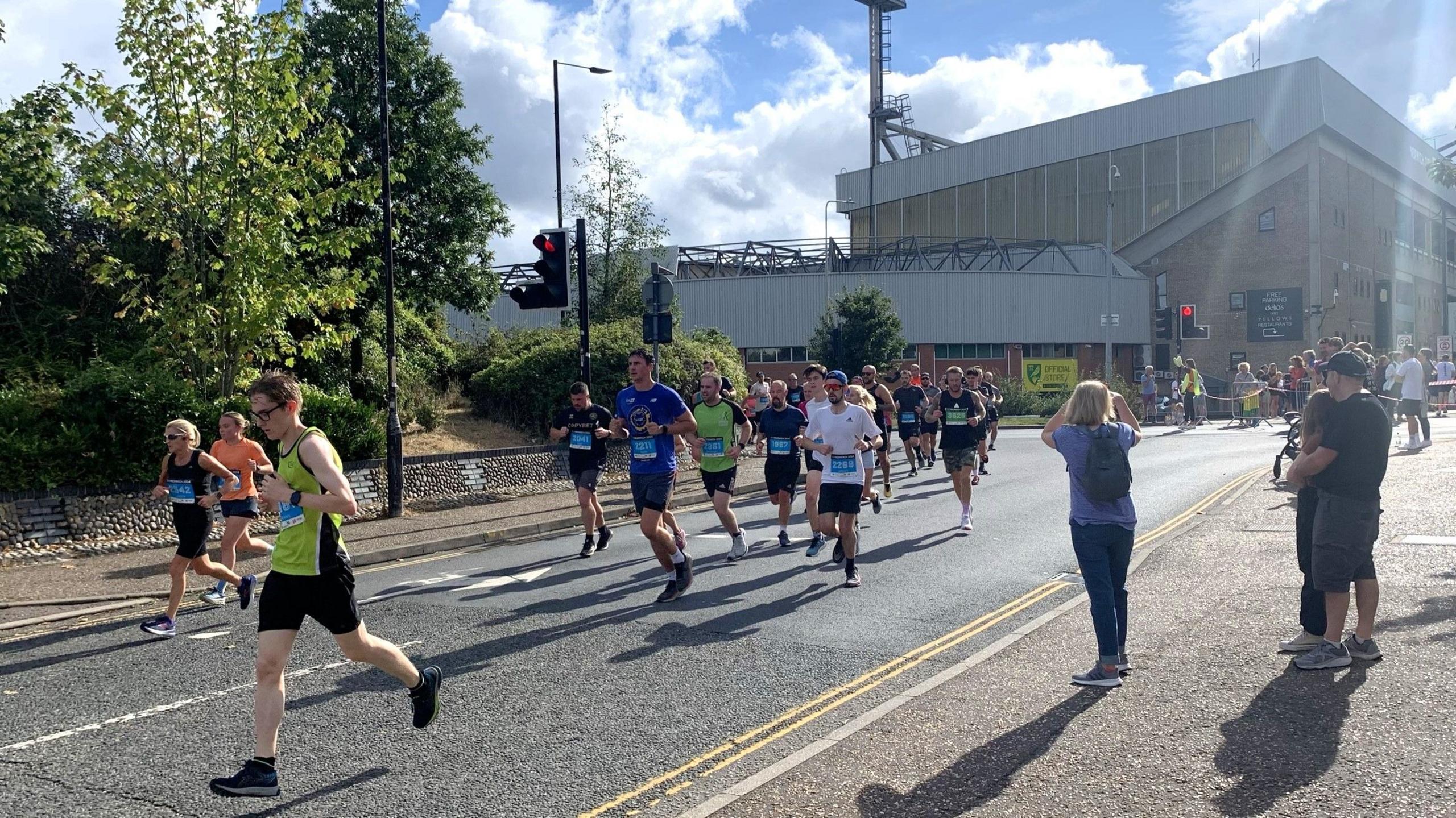 Runners dressed in various colours passing Carrow Road football ground. Spectators line the right hand side of the street. 