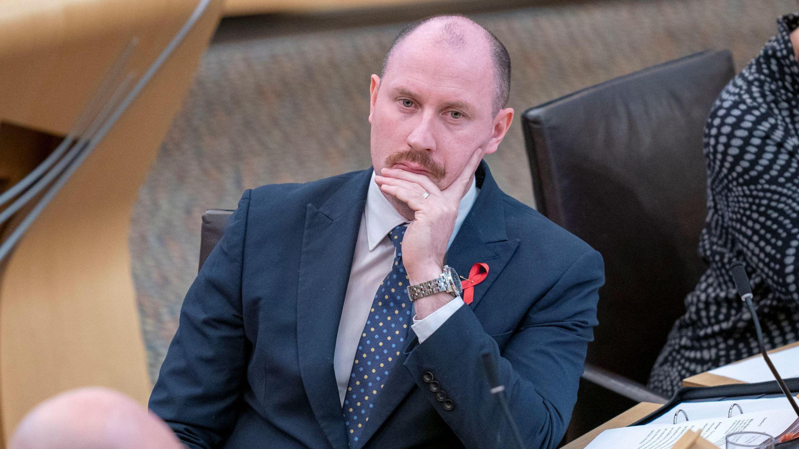 Neil Gray in the Holyrood chamber. He is resting on his chin in his hand and is looking thoughtful. He is dressed in a blue suit with tie, and has a moustache.