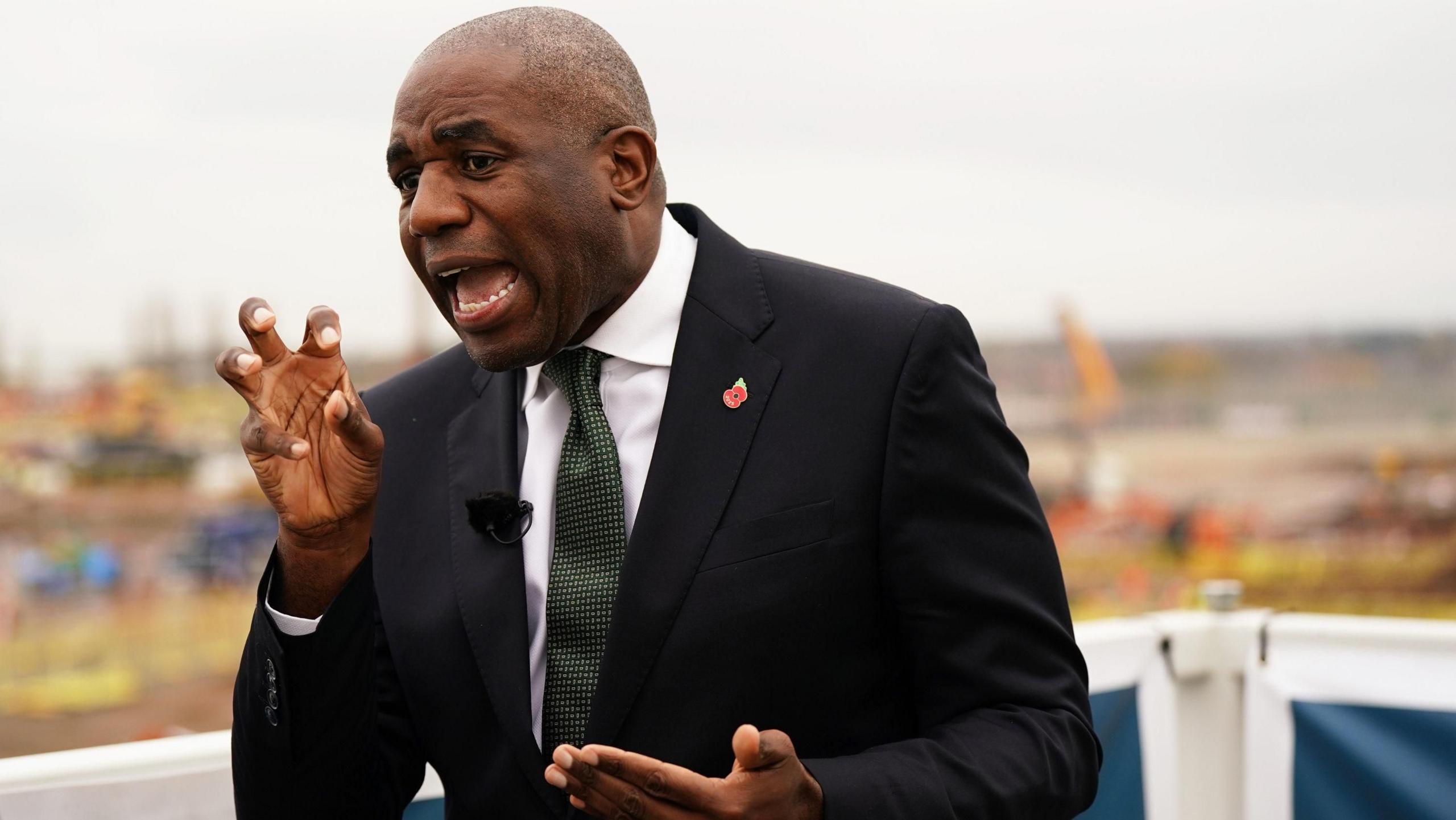David Lammy wearing a black suit jacket, white shirt, emerald tie, and a red figure-eight badge pinned on his jacket lapel, speaking to the media in front of a construction site 