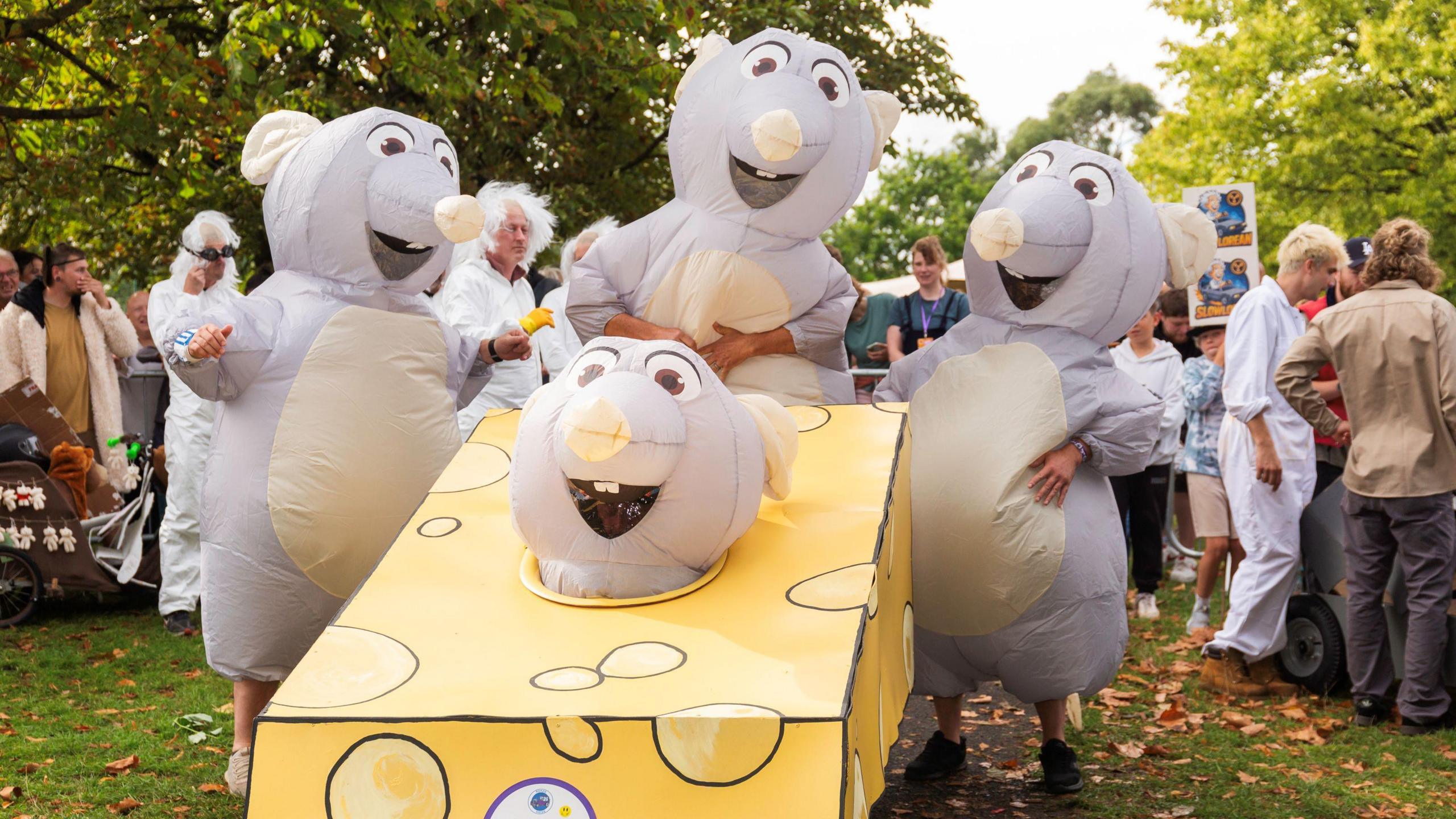 Four people in inflatable grey mouse costumes are seen with a home-made kart which is shaped to look like a piece of cheese. One of the mice is sitting inside the kart so only their head is visible. Behind them some spectators can be seen milling about at the event, the Krazy Races, in Victoria Park in Bristol.