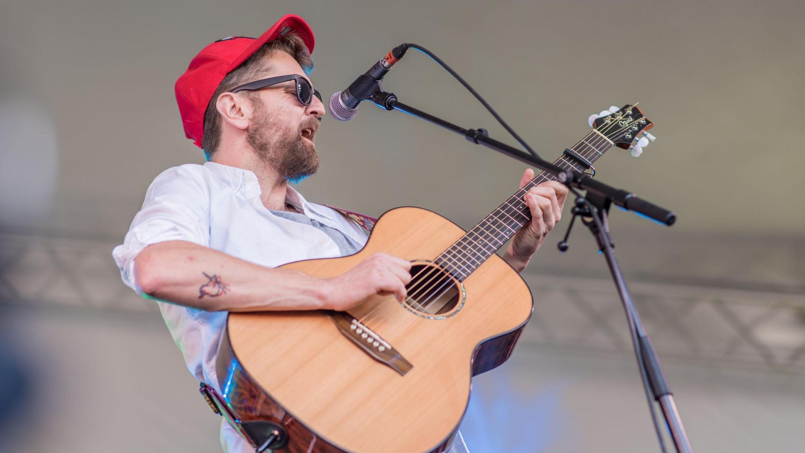 Ross Wilson playing onstage - he is strumming an acoustic guitar while singing, and is wearing a red baseball cap, sunglasses and white shirt