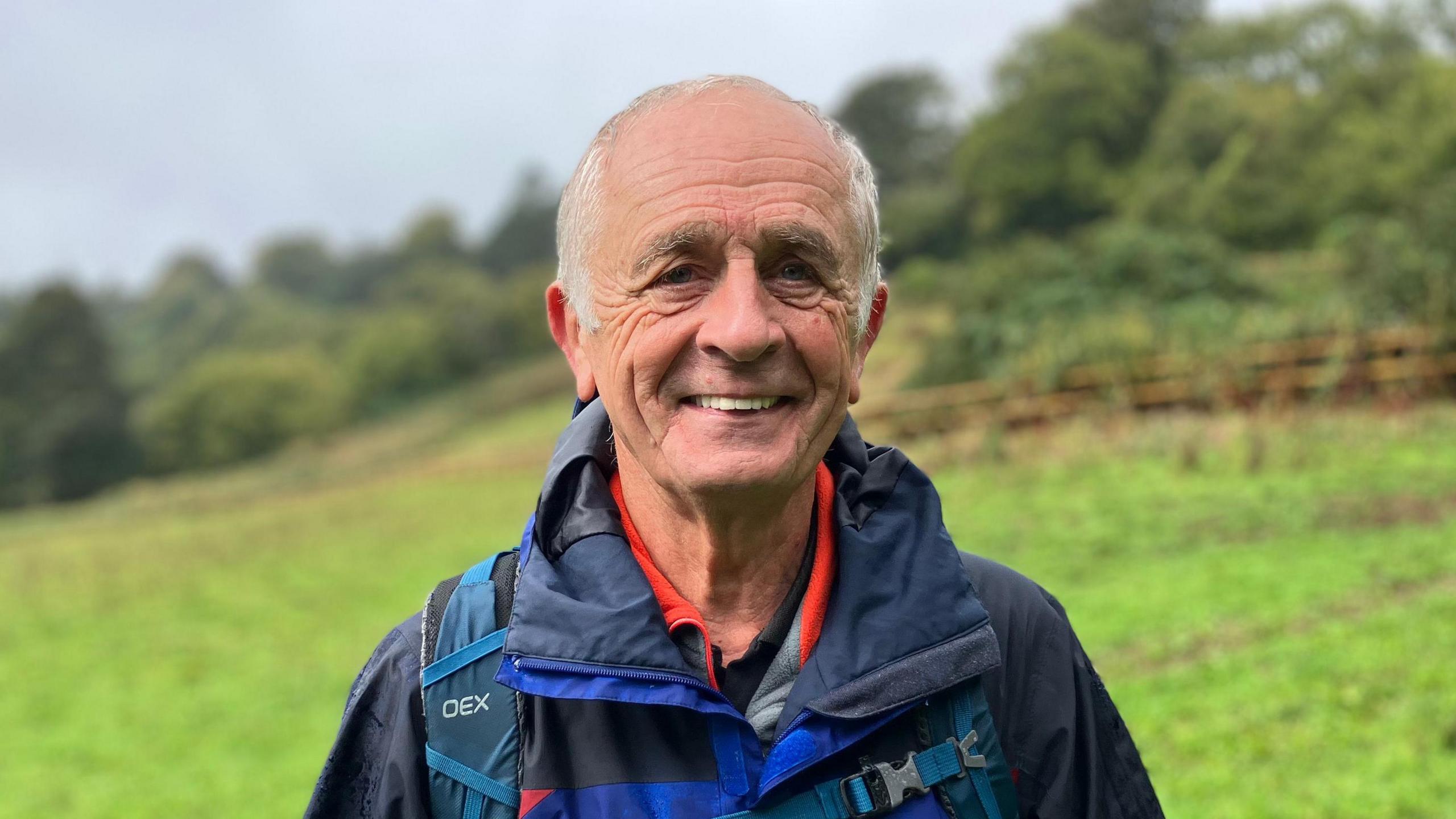 Man in a blue and black coat with a rucksack. He has white/grey hair and is smiling