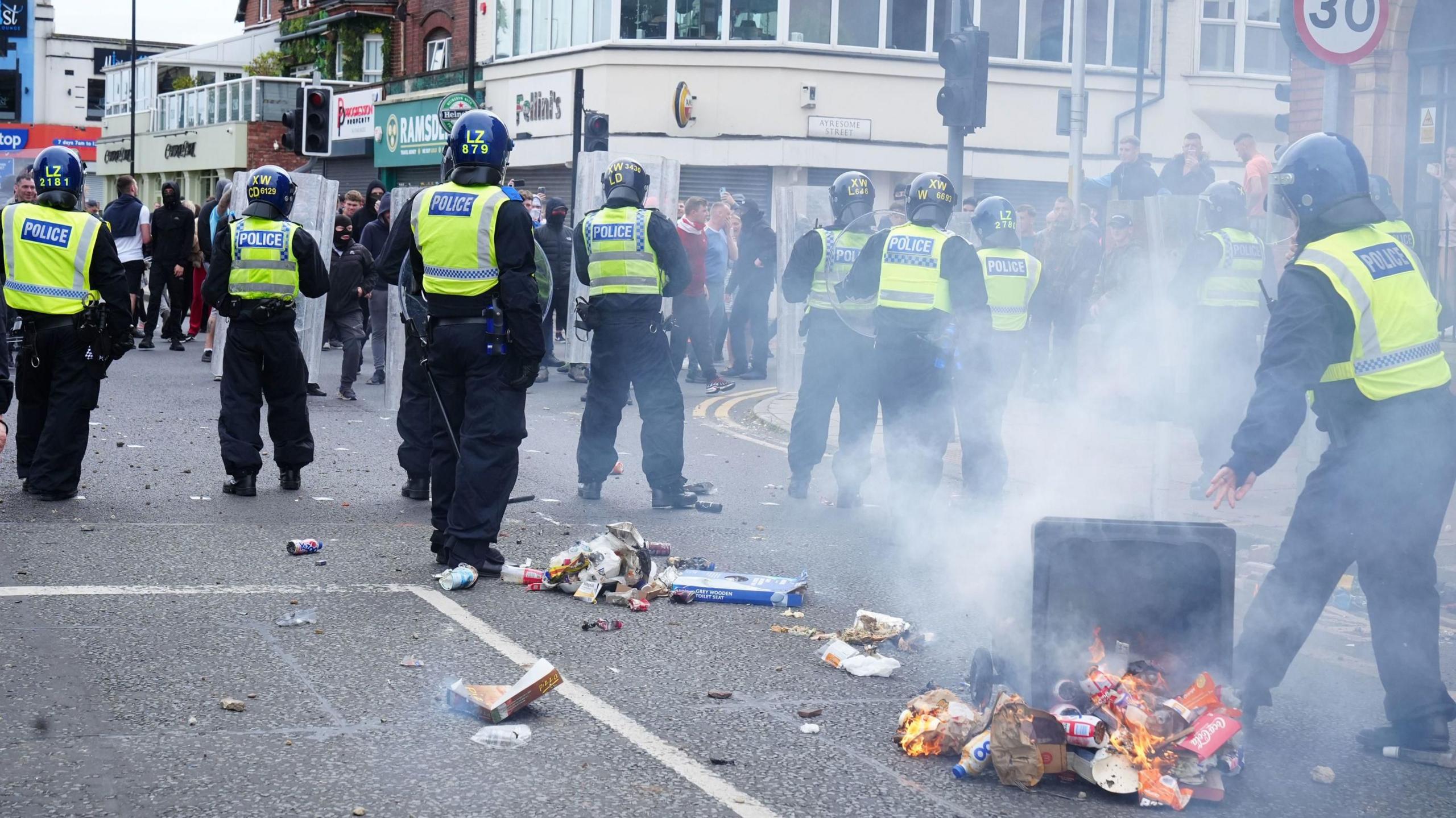 A number of police officers wearing protective gear and carrying shields face rioters in a Middlesbrough street. In the foreground, smoke is rising from an overturned wheelie bin which has been set on fire. Items of litter are spilling out of the bin on to the street.