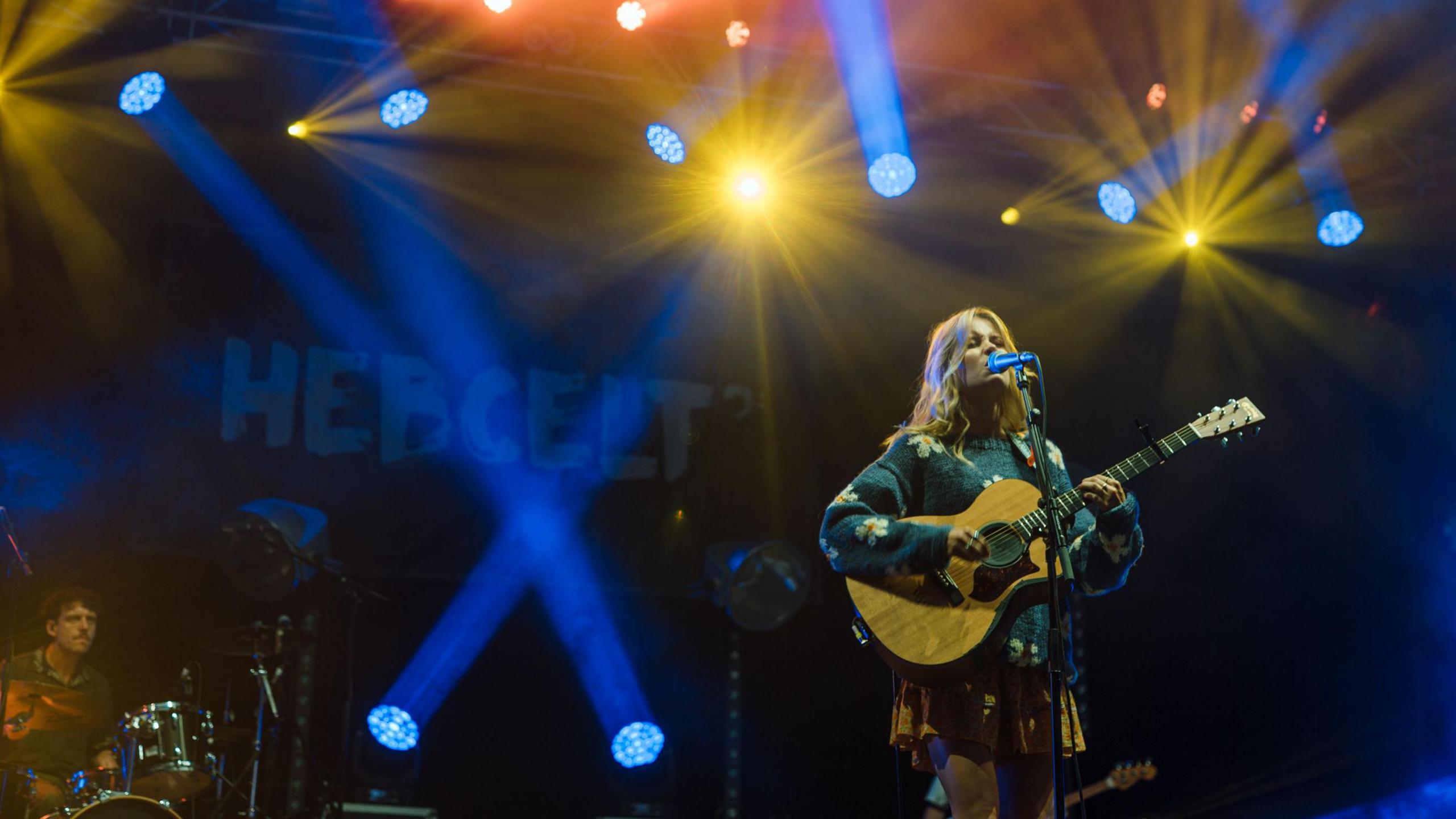 Woman with long, blonde hair sings into a mic as she plays guitar. The stage is lit in gold and blue lights and there is a drummer behind her