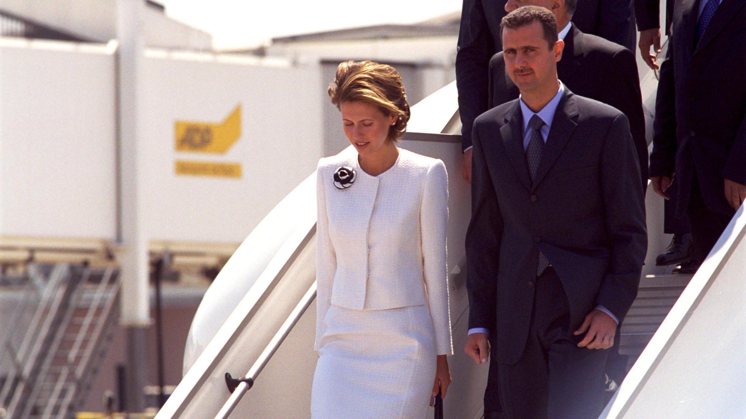 Asma al-Assad in white and Bashar al-Assad in black descend a plane staircase.