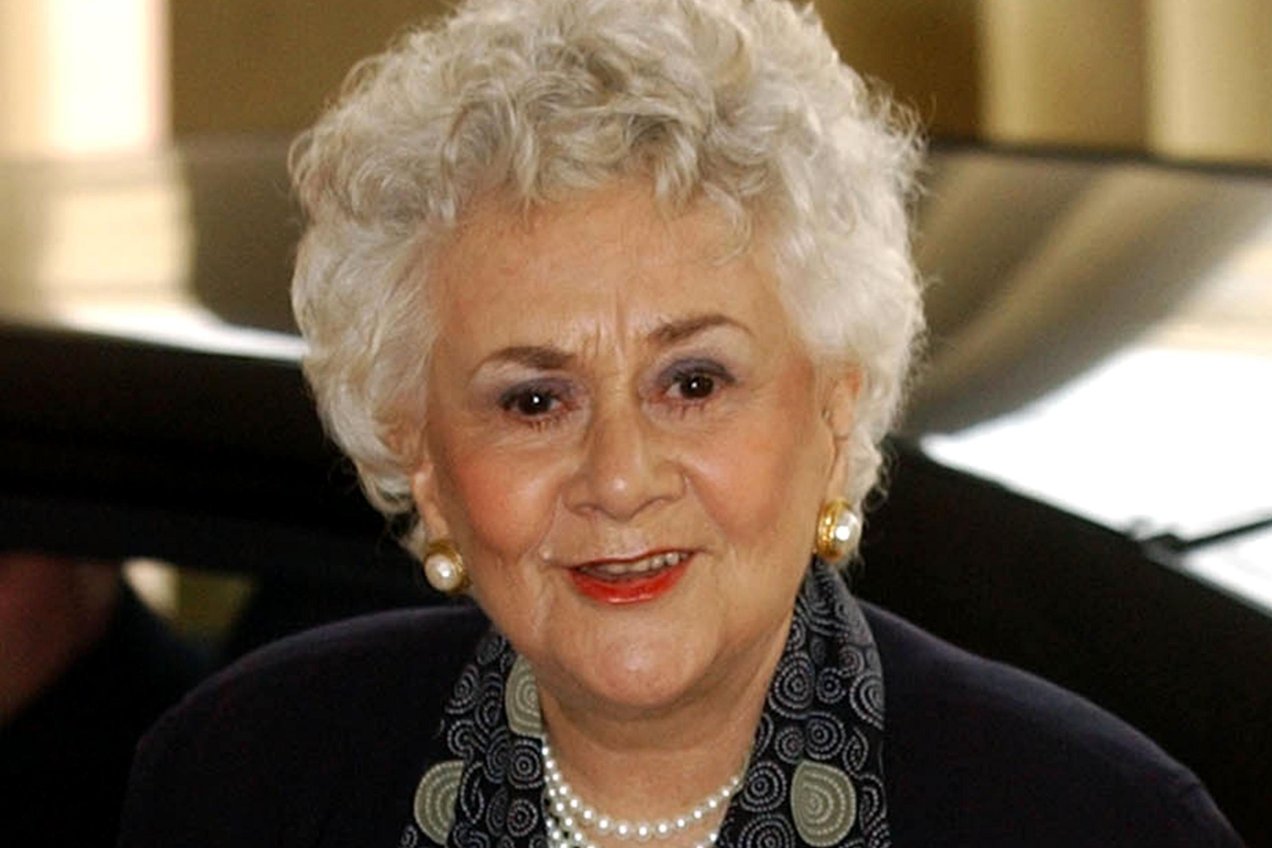 Dame Joan Plowright ka flokë të shkurtër e kaçurrela të bardhë dhe mban veshur një veshje të errët të kombinuar me një shall me motive dhe një gjerdan me perla. Vathët e artë janë të dukshëm, dhe sfondi tregon një pjesë të një automjeti dhe një ambient të brendshëm me ndriçim të ngrohtë.