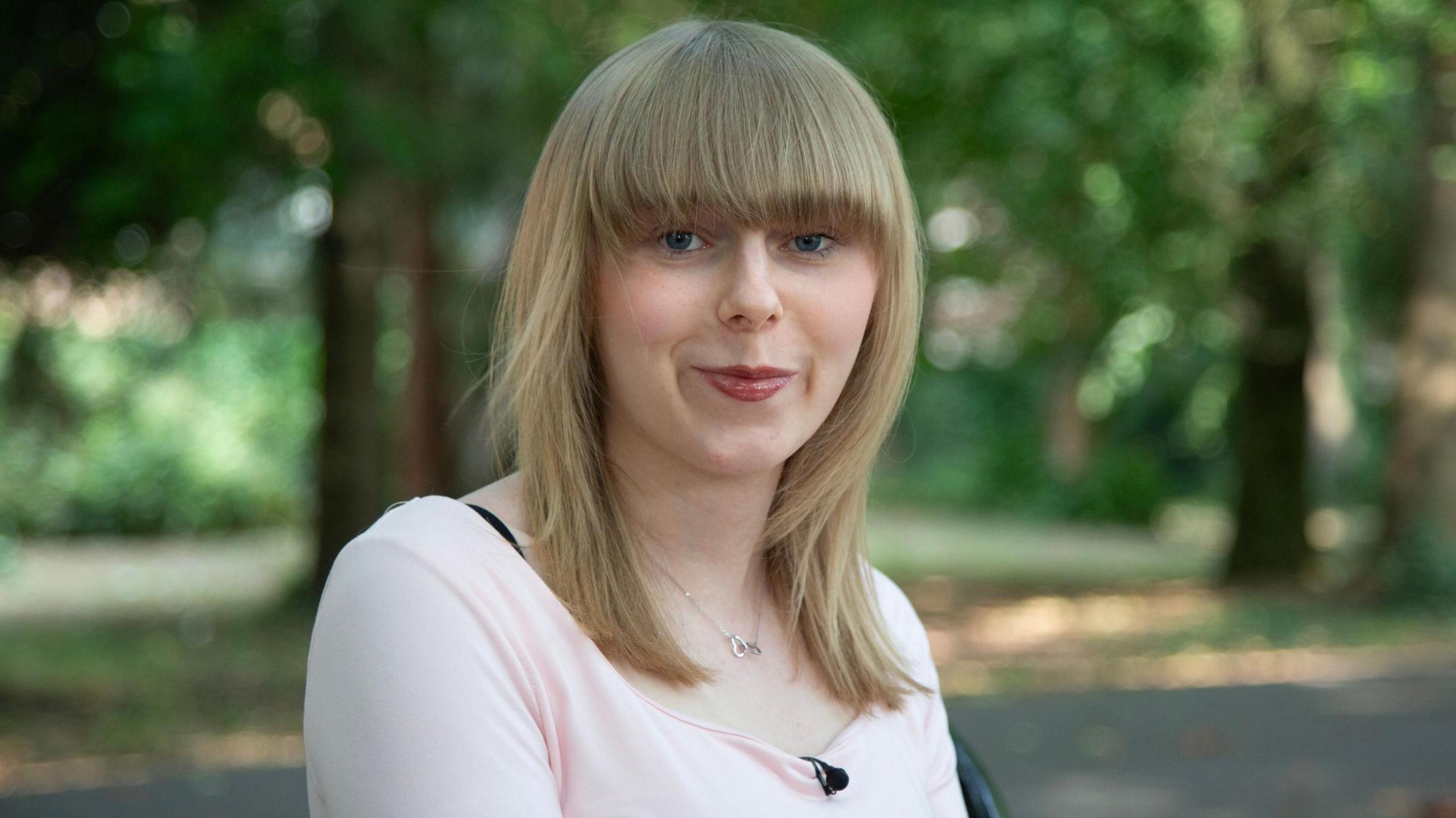 Emily pictured outdoors in a natural setting, wearing a pale pink top against a backdrop of softly blurred greenery.
She has shoulder length blonde hair with a fringe coming down to her blue eyes.