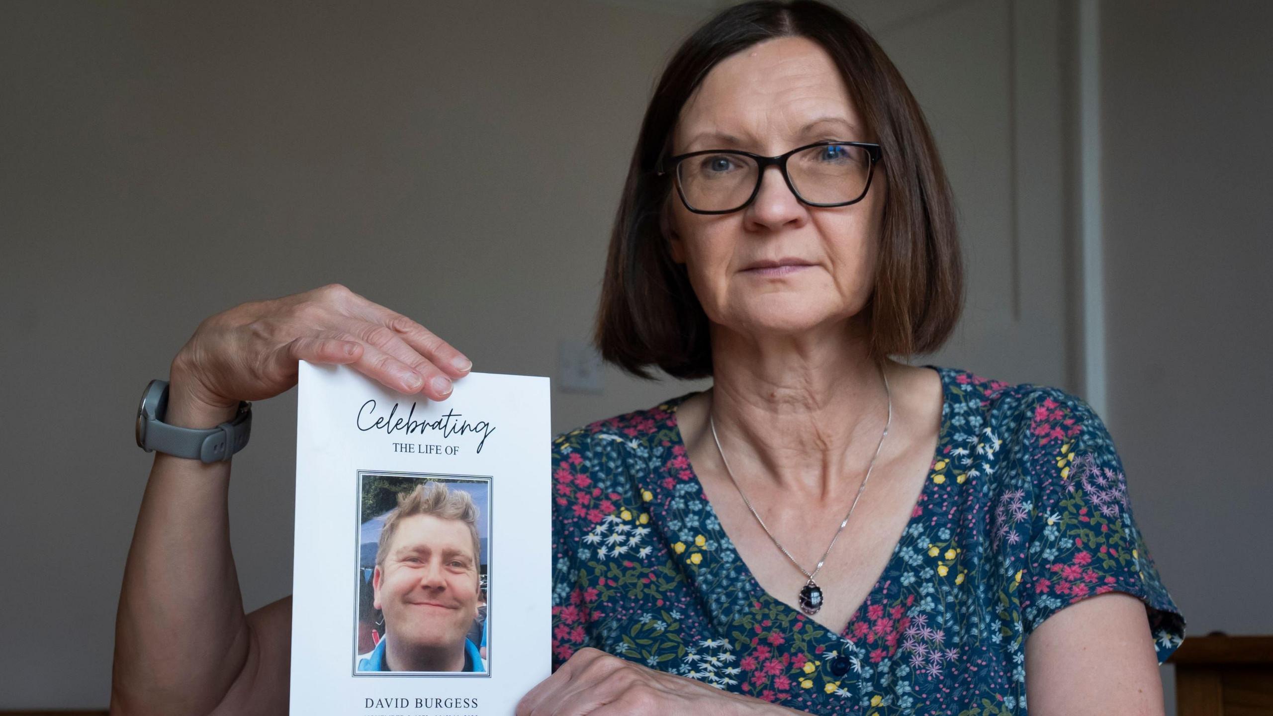 Sandra O'Hagan holding a funeral order of service for her late partner David Burgess. She is wearing a floral dress with different coloured flowers and a pendant necklace. She has bobbed brown hair and is wearing glasses. The order of service says 'Celebrating the life of David Burgess', with a photo of Mr Burgess smiling.