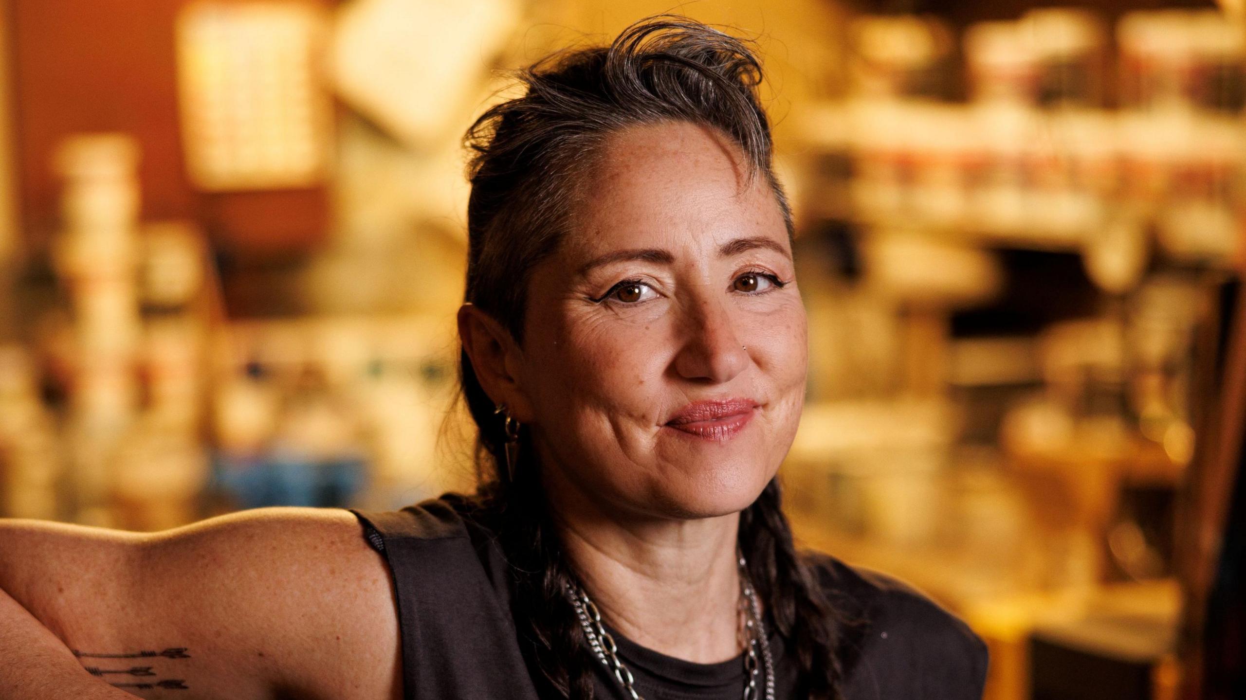 KT Tunstall in black T-shirt and necklaces, with her hair swept back off her face. She is smiling. The gold-coloured backdrop is out of focus.