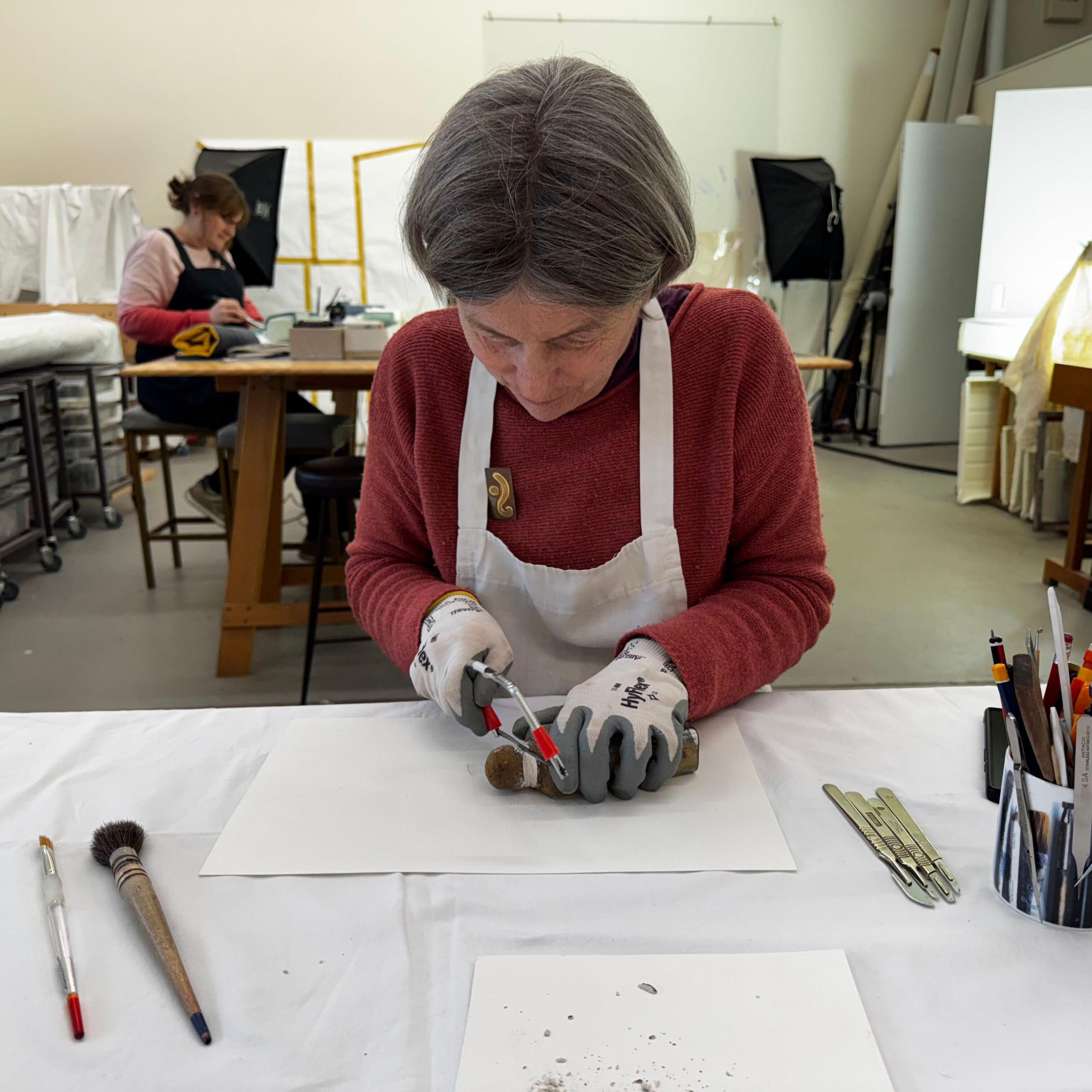 Helen Creasy has grey bobbed hair. She is wearing a maroon top and white apron. She is also wearing gloves and is cutting the bottle with a small saw.