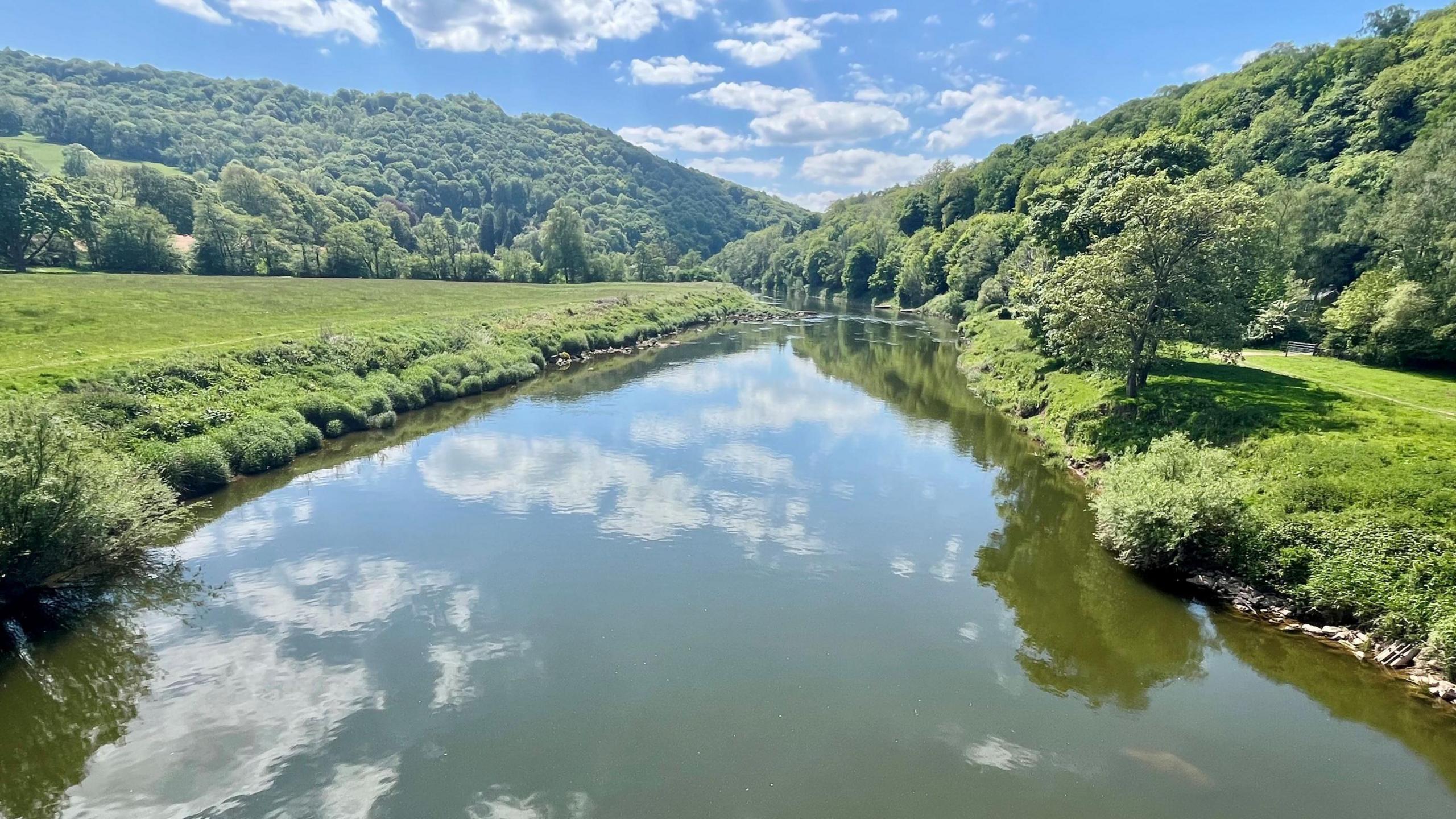 The River Wye is pictured on a sunny day, with banks of green trees either side. The water is a mixture of greeny/grey and the sky is largely blue with some white clouds