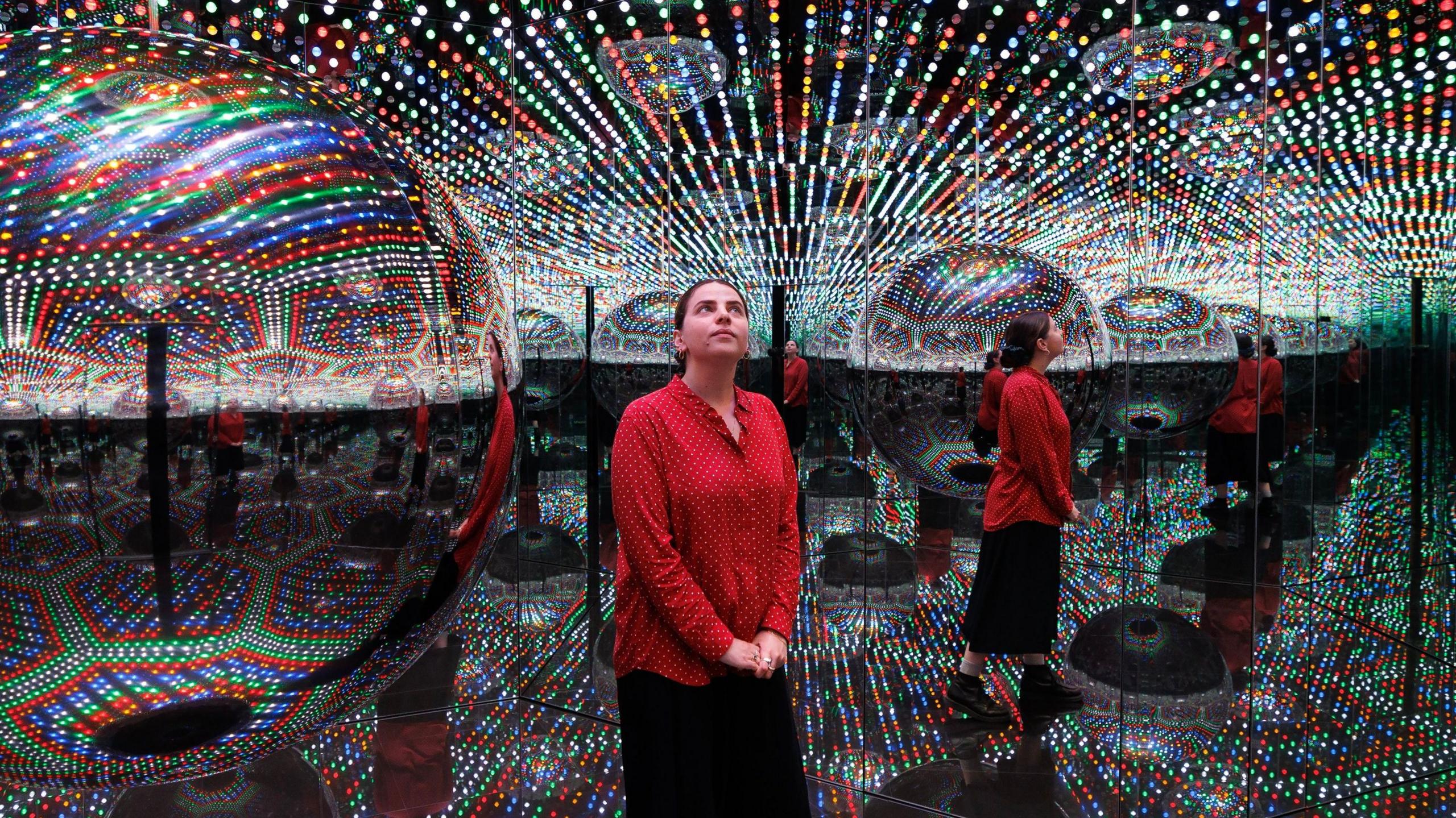 A visitor standing in one of Kusama's Infinity Rooms, seen reflected several times in mirrors surrounded by lights.