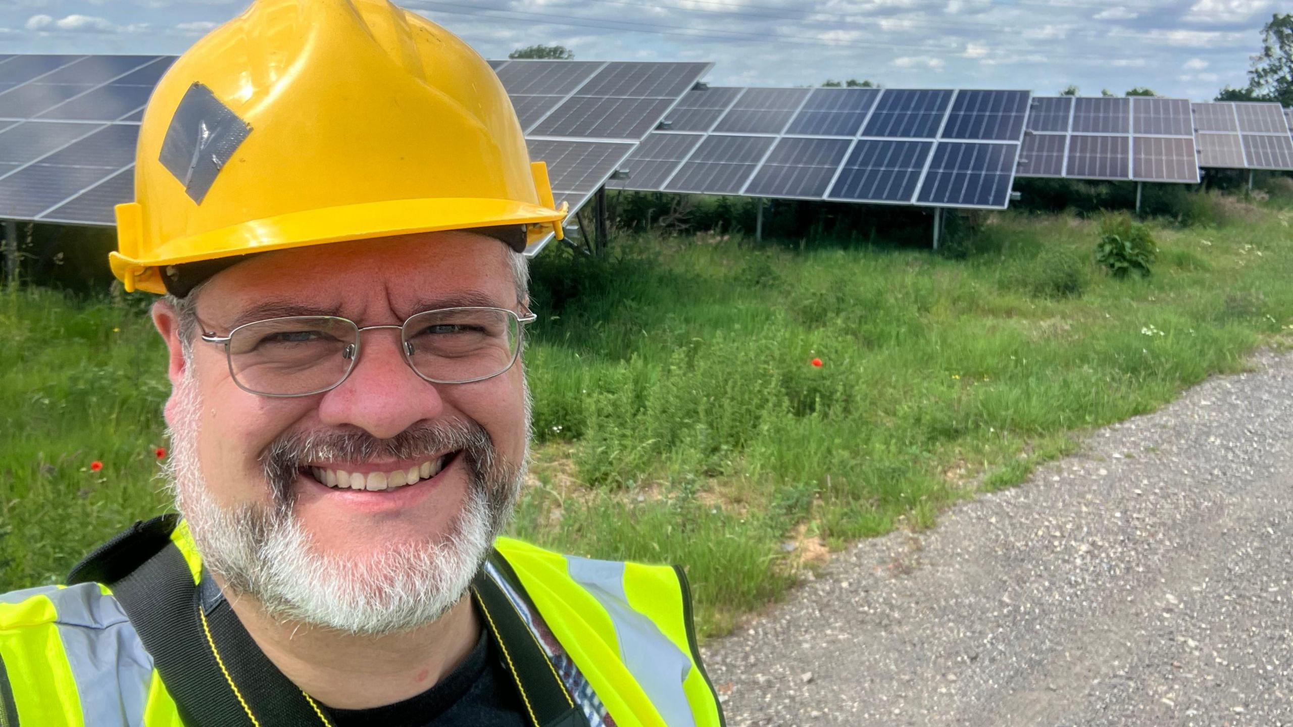 Gareth Simkins, smiling, bearded, glasses, yellow hard hat and high vis top smiling at camera in front of rows of ground mounted solar photovoltaic panels. Poppies and other wild flowers are visible in front of the panels.