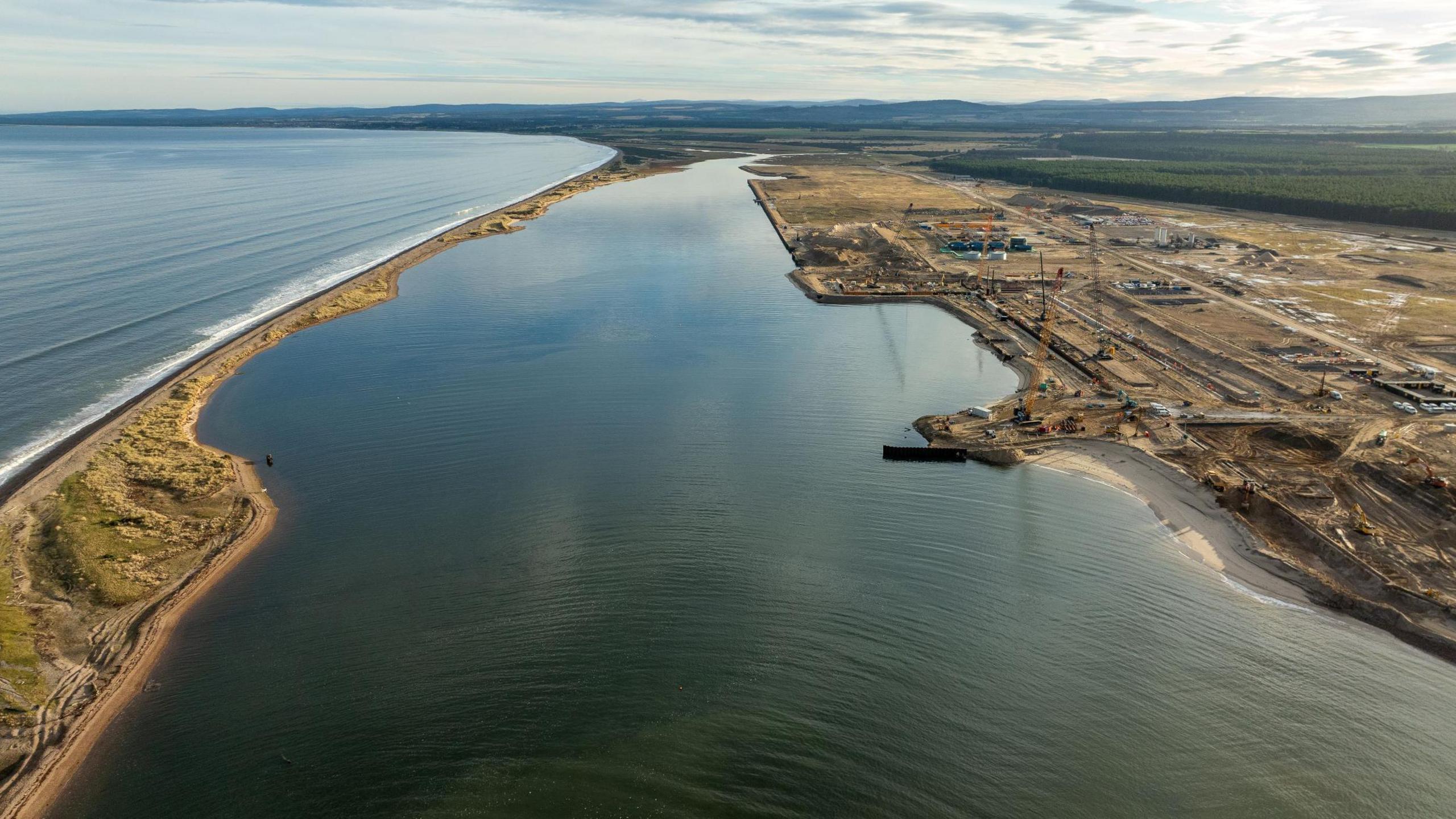 The port is a vast brownfield site on the Moray Firth. Construction equipment, including cranes, are just visible and a long spit of sand stretches towards the camera from a distant shoreline.
