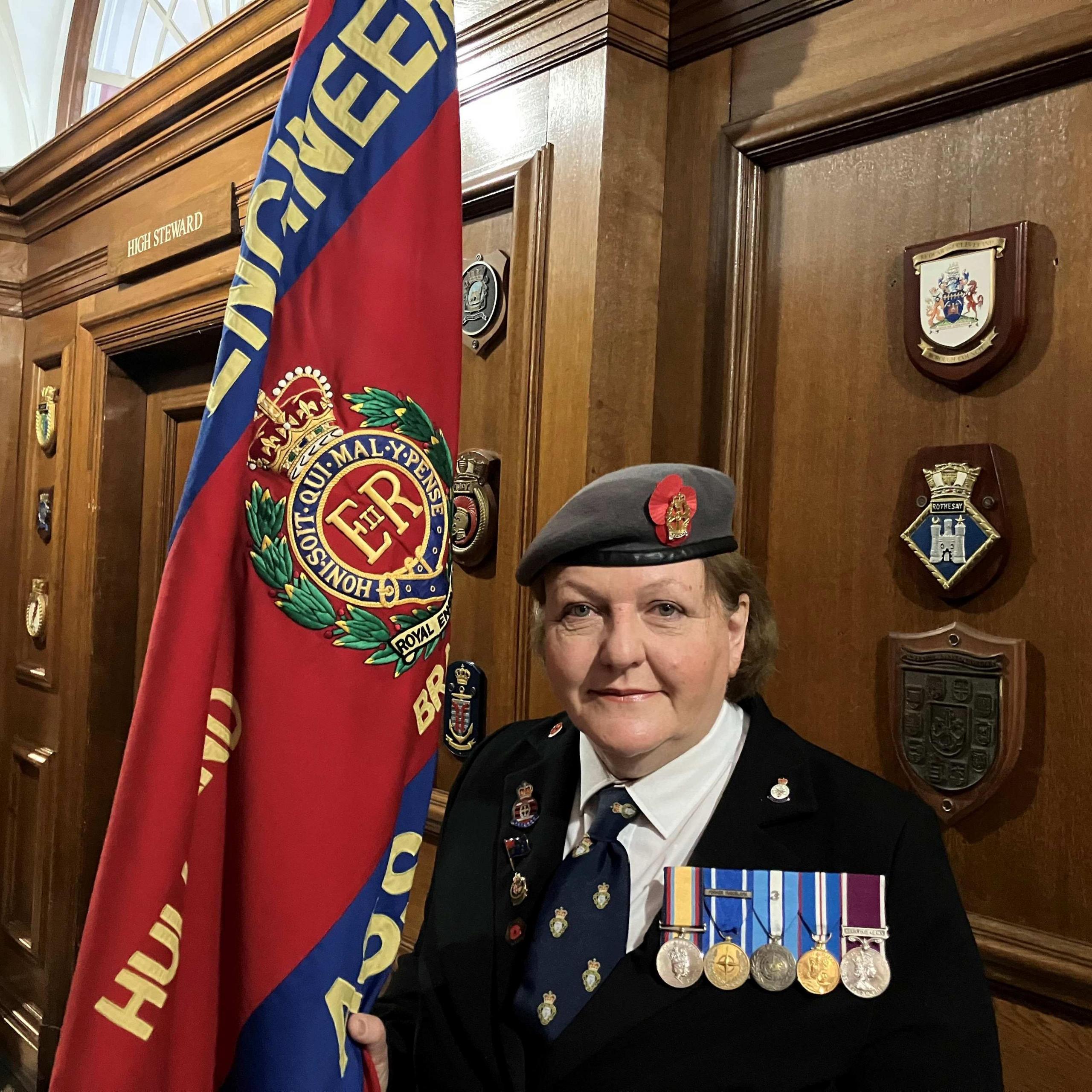 A woman wearing a black blazer, regimental beret, tie and a row of medals stands in a grand-looking, wood-panelled corridor while holding the flag of a regimental association, which is coloured scarlet and blue and includes a regimental badge.