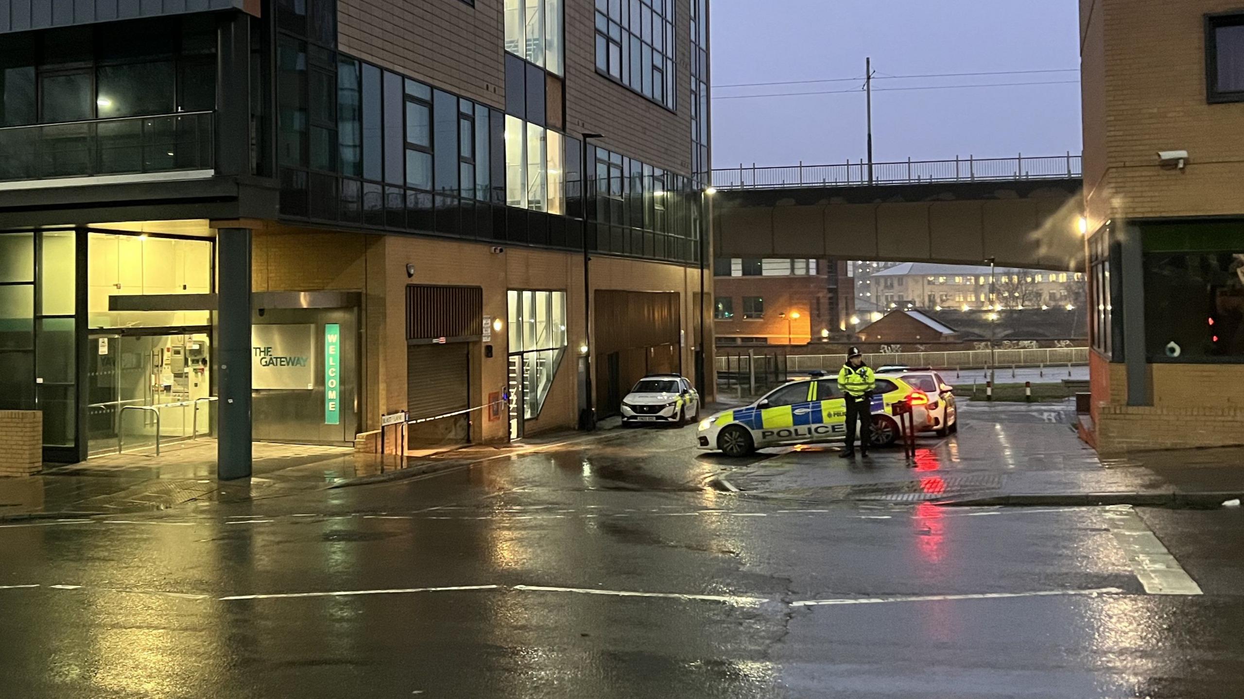 A police officer standing by a police car, just next to the Gateway apartment block in Sheffield. There is some police tape across the main entrance.