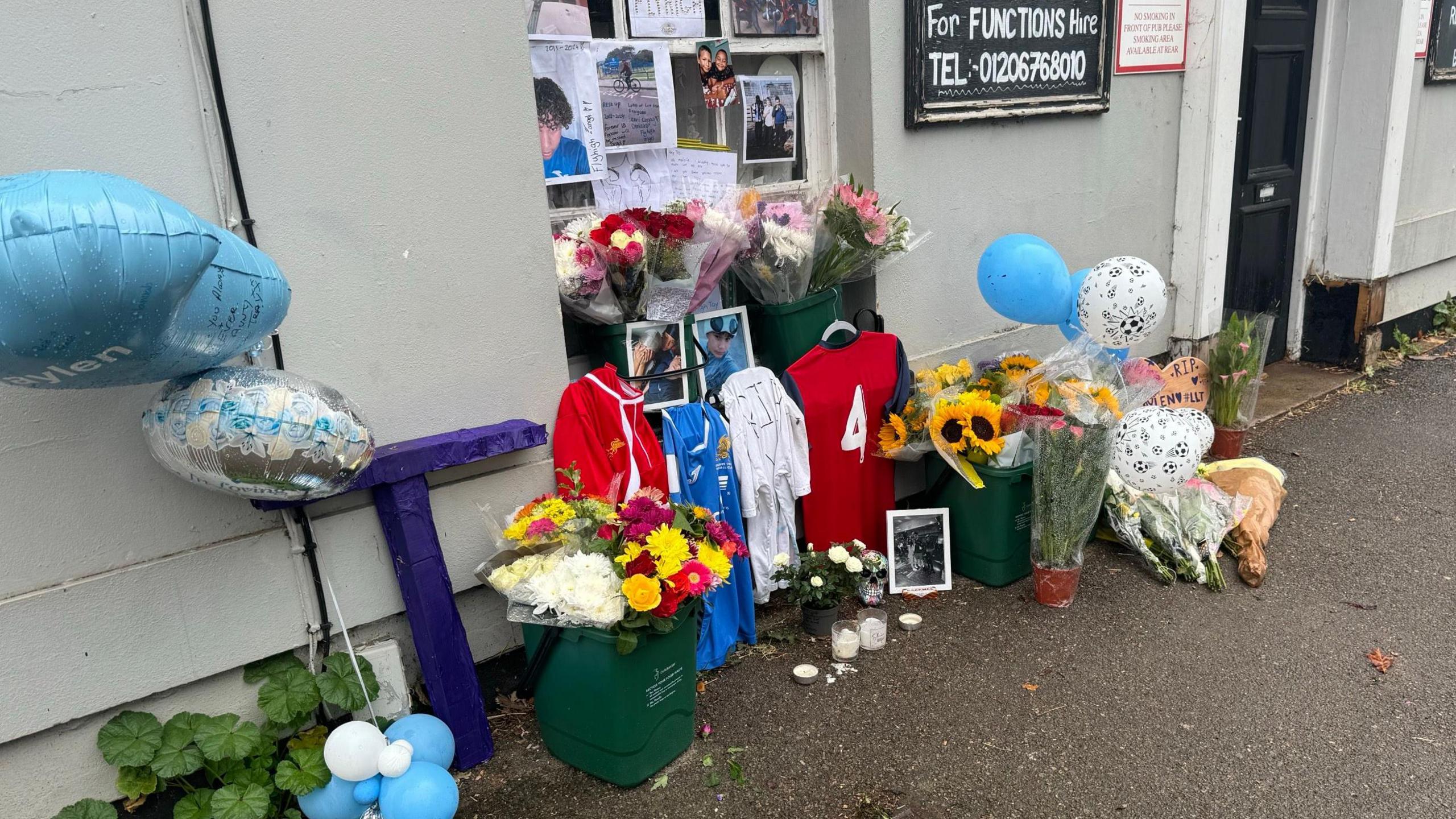 Floral tributes, balloons, football shirts, and photographs outside a pub.