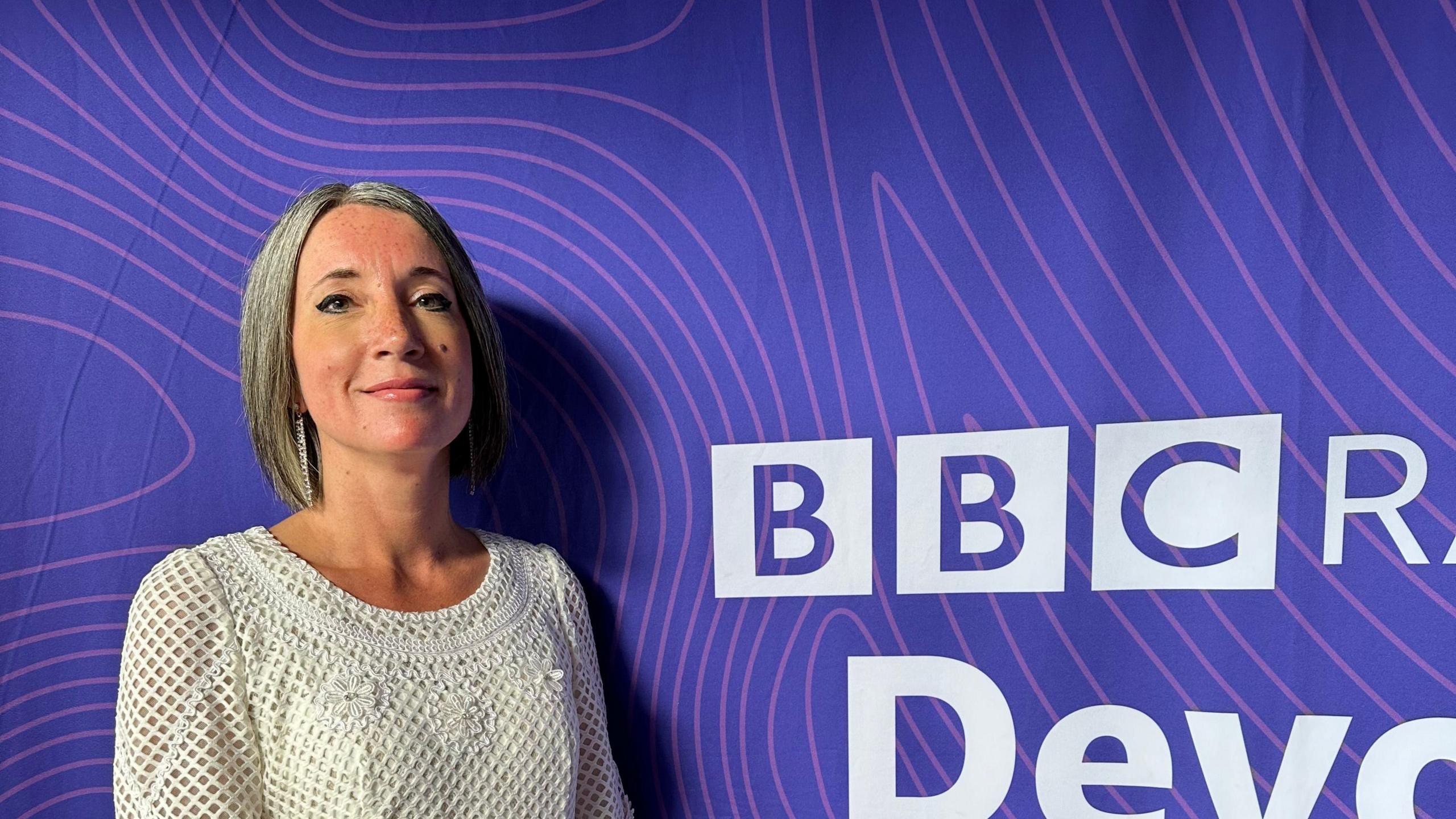 Clare Matheson wearing a white dress with long silver earrings. She stood against a purple background with the words 'BBC' on it.