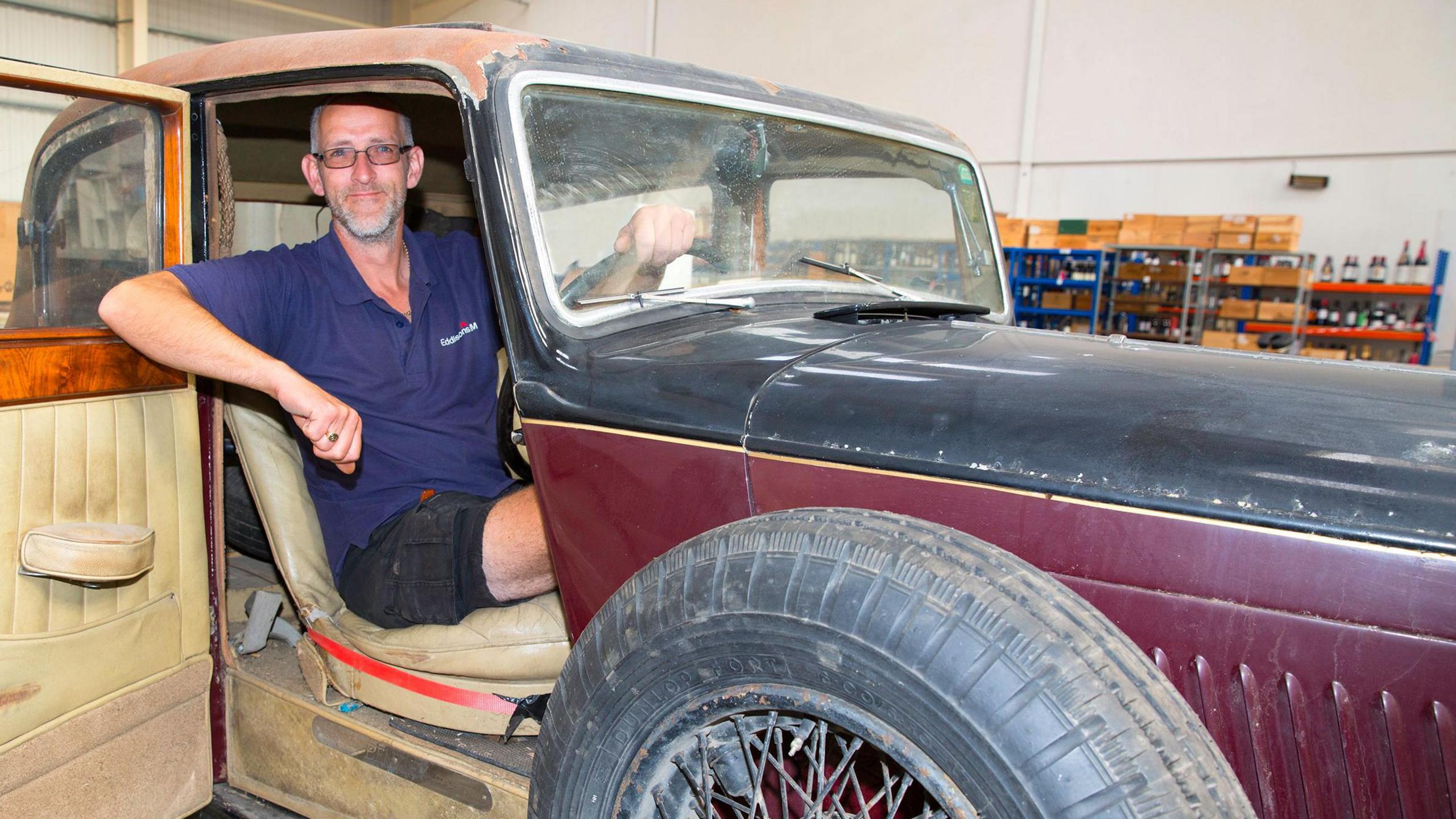 A man sits in the driver's seat of the Bentley, which has a wood and cream leather interior but is in need of restoration